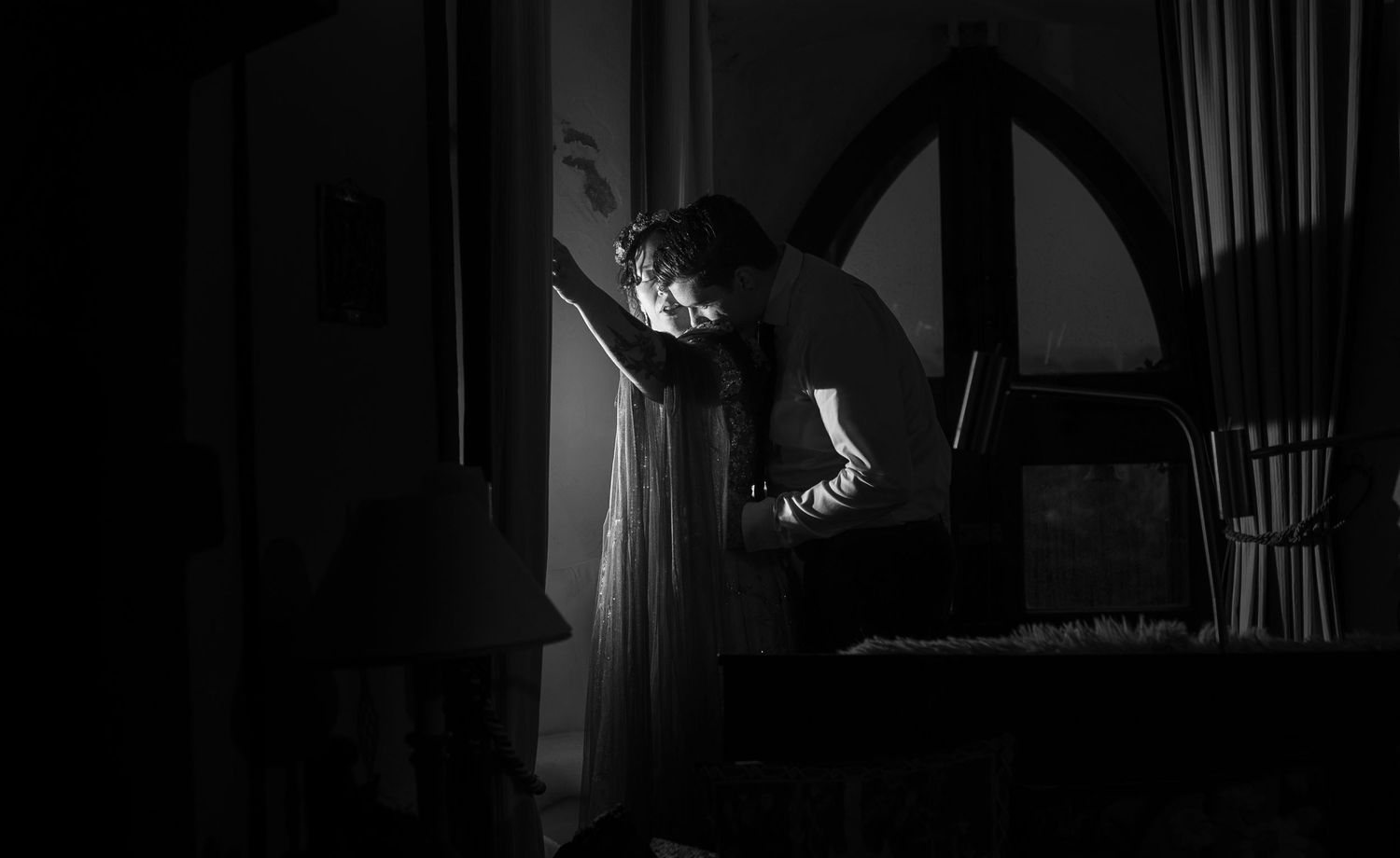Black and white photo of a person in white gown standing by arched window in dimly lit room.