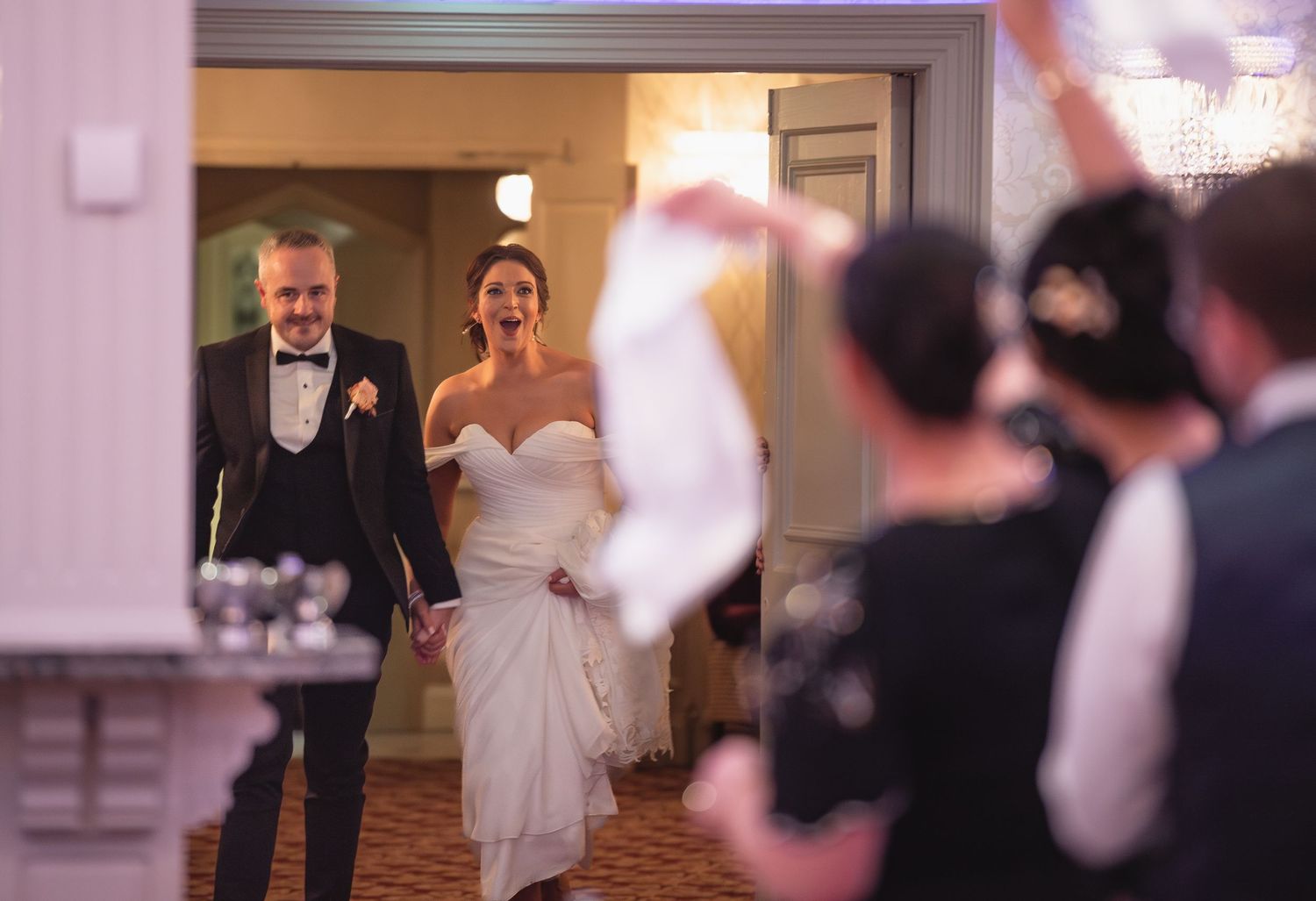 Joyful bride and groom make grand entrance at elegant wedding reception while guests celebrate with waving napkins.