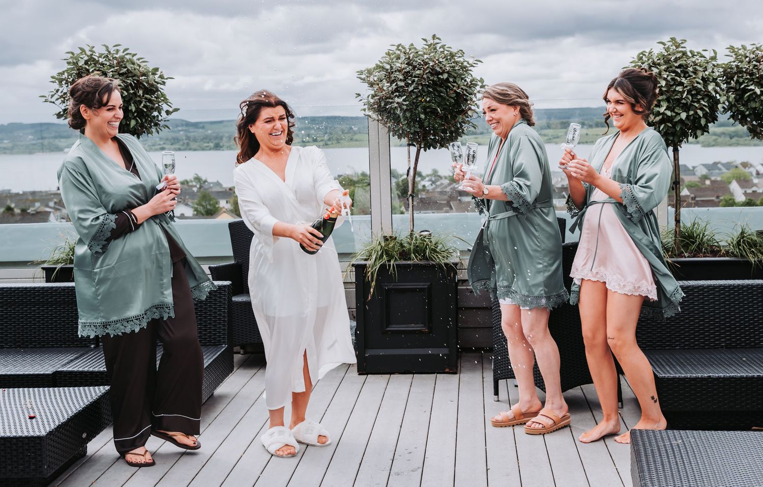 Bride in white robe pops champagne with bridesmaids in sage green robes on a rooftop deck with scenic water views.