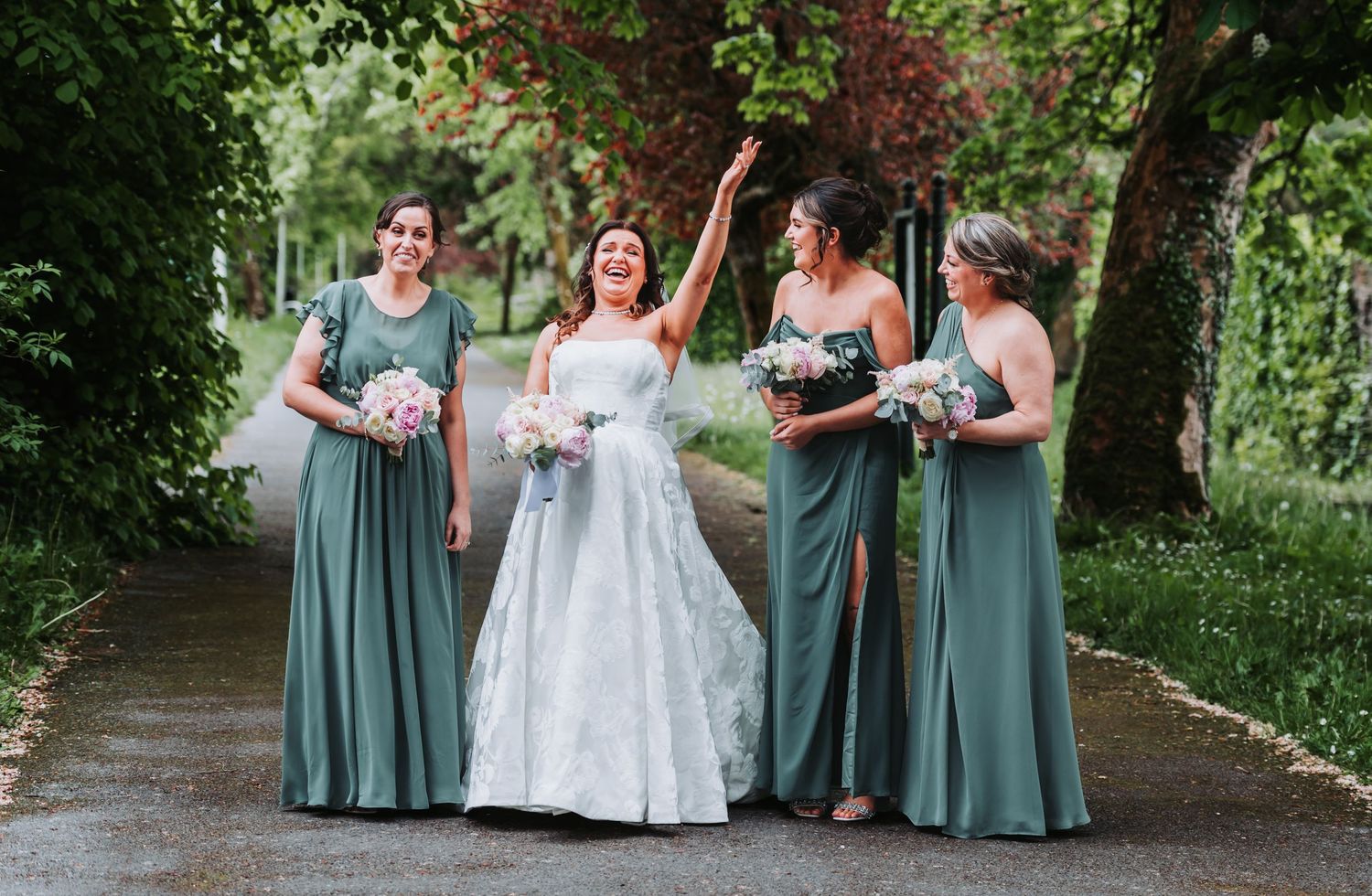 Bride in white gown laughing with three bridesmaids in sage green dresses, holding bouquets outdoors.