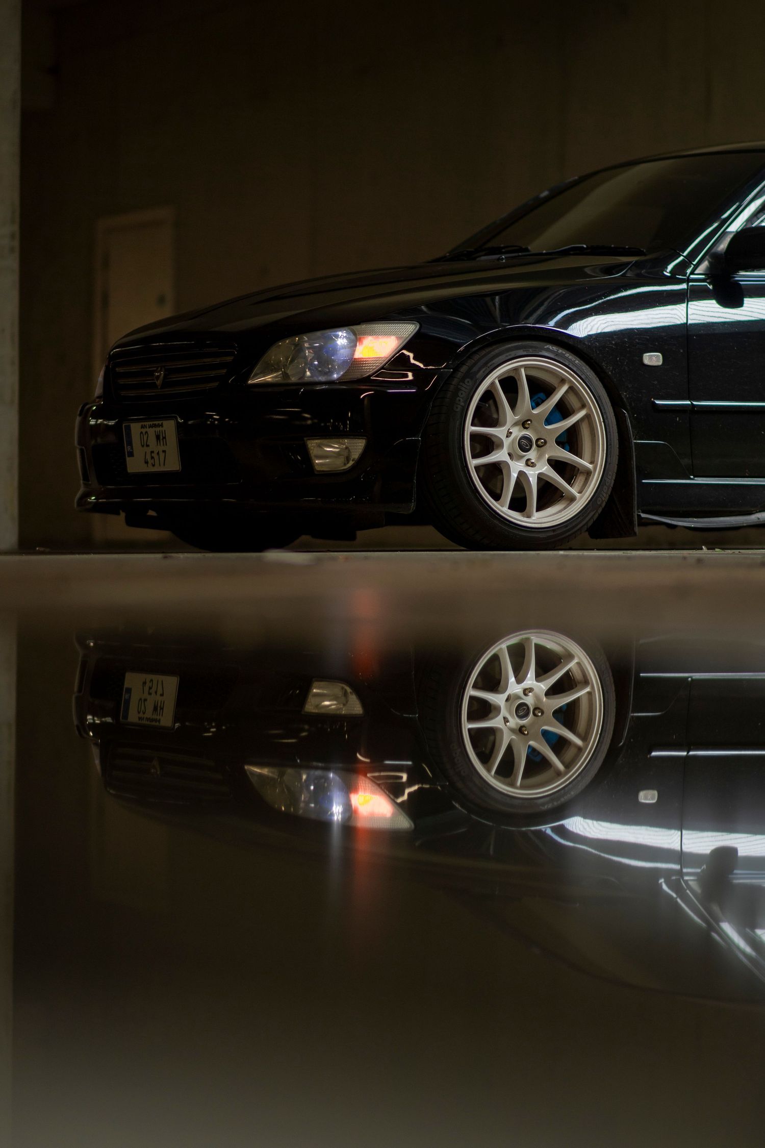 Black sports car with silver alloy wheels reflected on glossy garage floor in dramatic low-light photography.