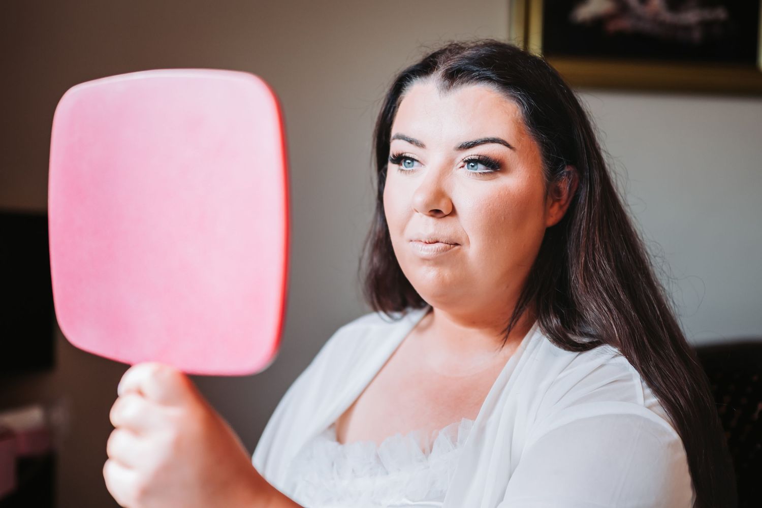 Bride checking her makeup in a pink hand mirror while getting ready on her wedding day.