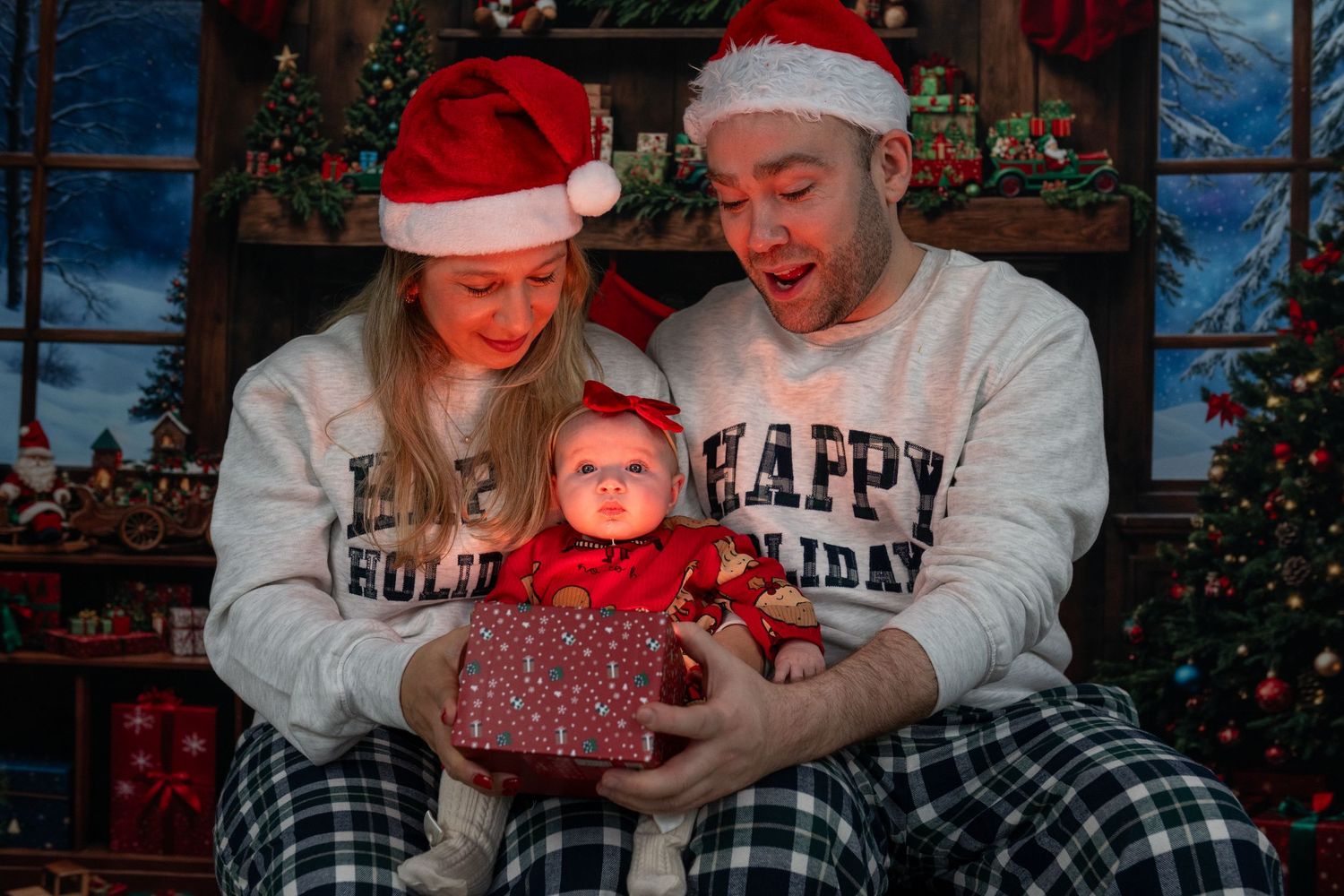 Family in matching holiday pajamas and Santa hats holding baby in red outfit with Christmas gift.