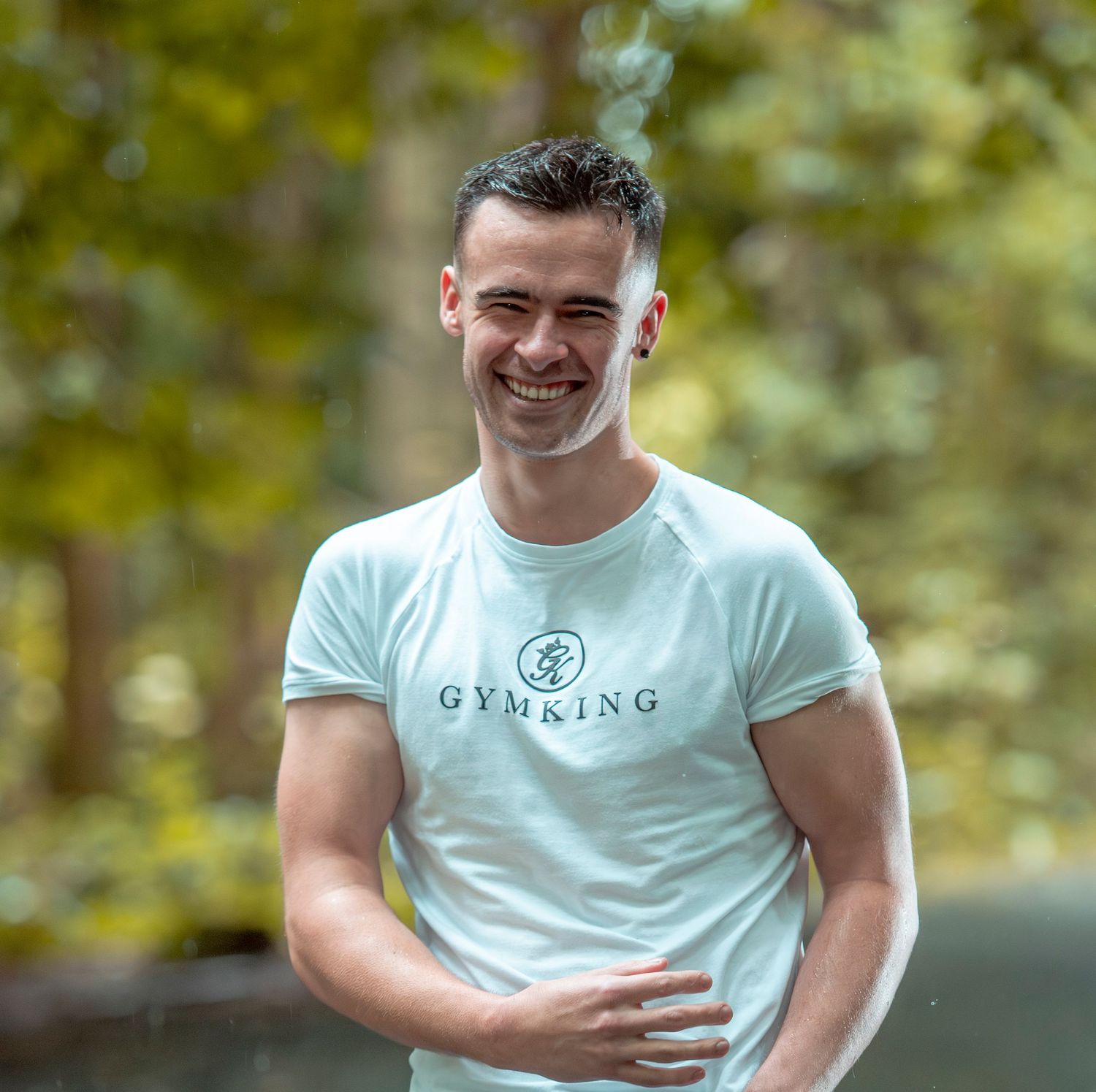 Smiling young man wearing a light blue Gym King t-shirt outdoors with green trees in the background.