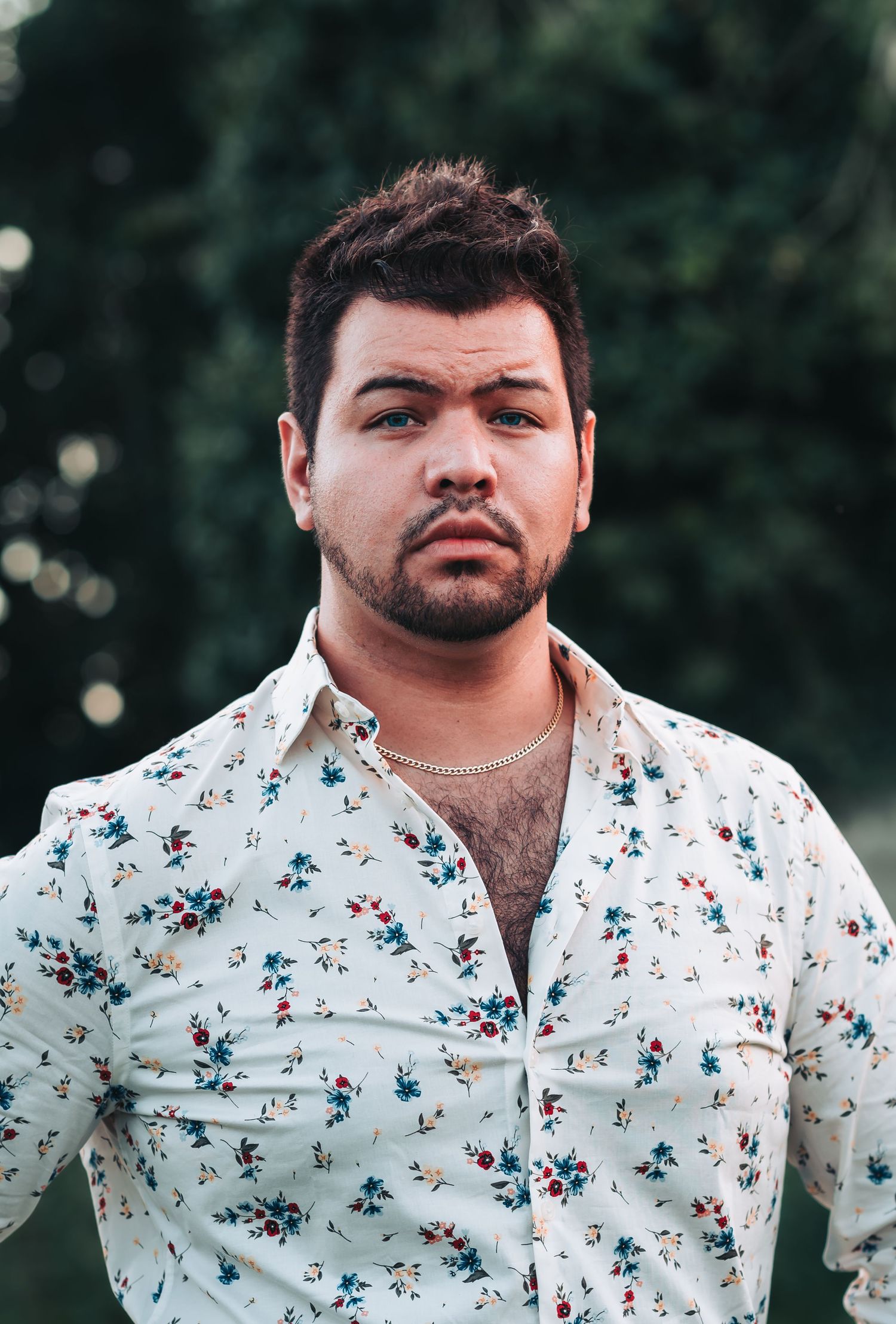 Young man with dark hair wearing a floral shirt and gold chain necklace, posing outdoors with green trees behind him.