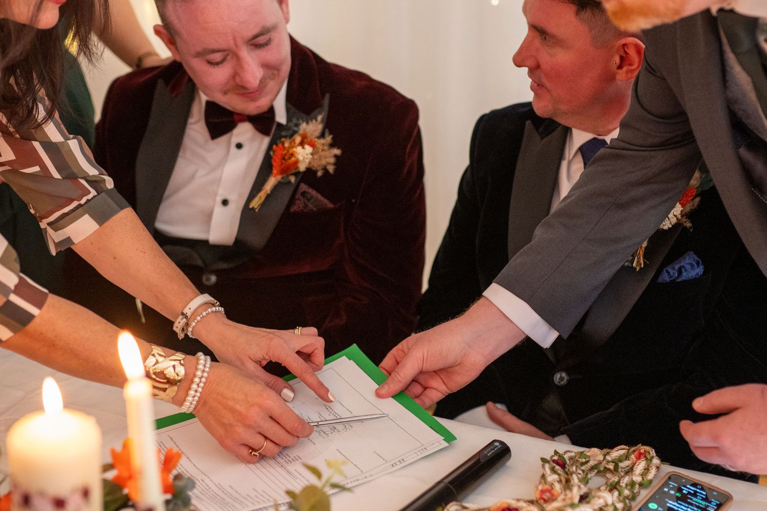 Two grooms signing their wedding certificate by candlelight, one wearing a burgundy velvet tuxedo.