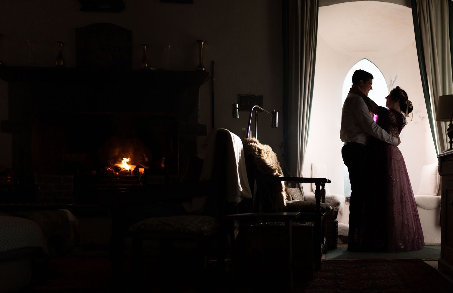 Couple silhouetted by arched window embrace romantically beside glowing fireplace in dark castle room.