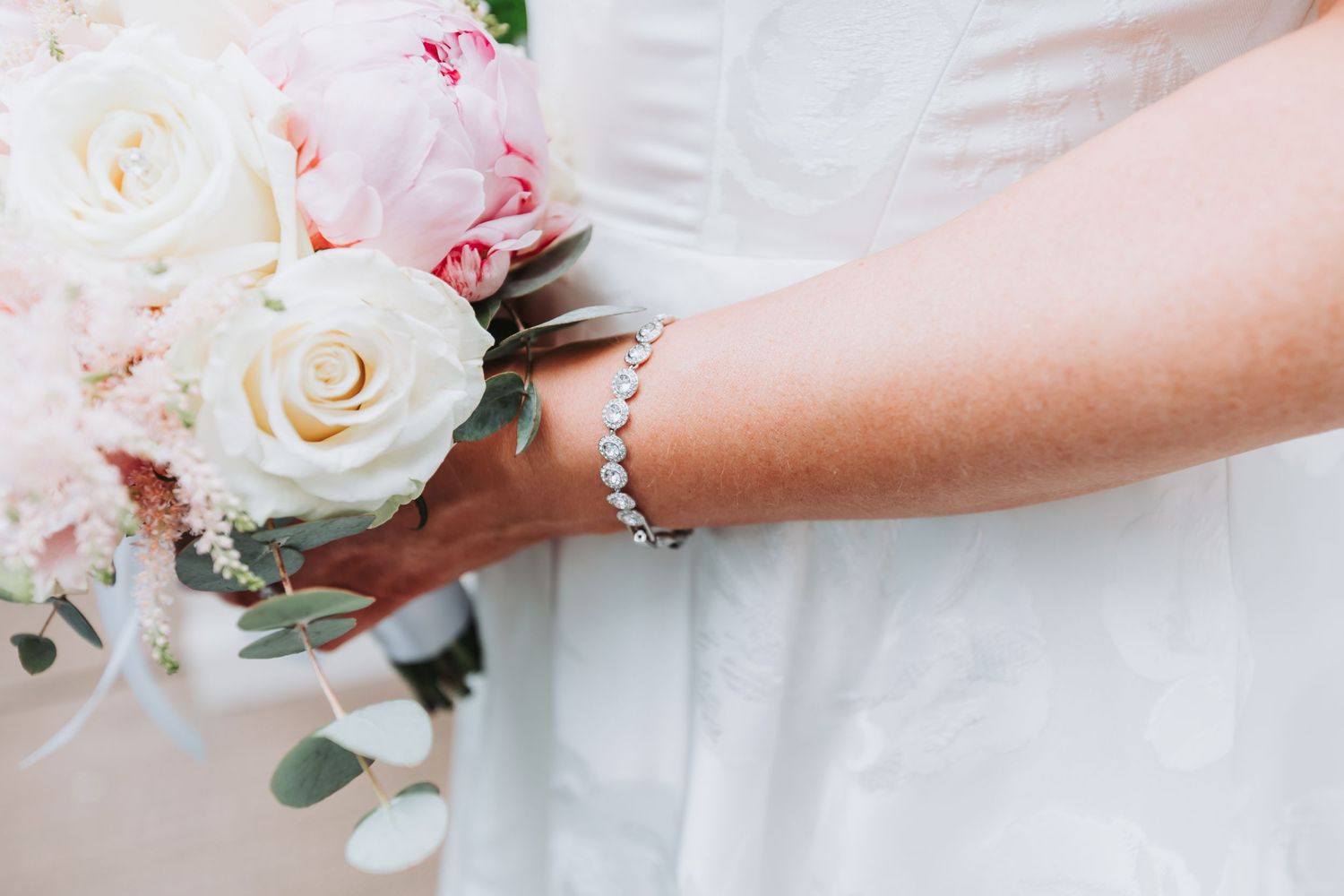 Bride holding a pink and white rose bouquet, wearing a delicate diamond tennis bracelet on her wrist.