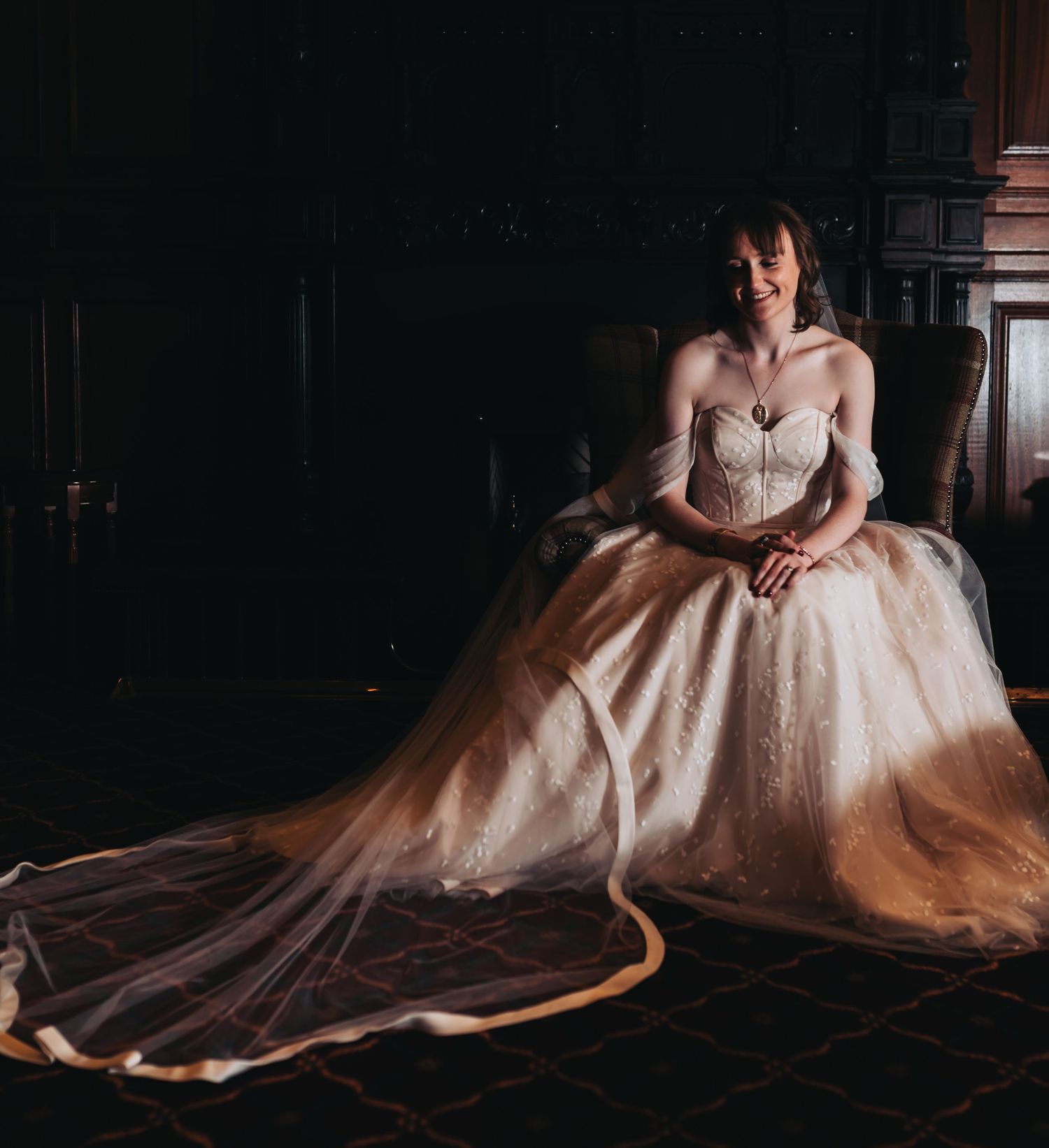 Bride in strapless lace ballgown with long train sits smiling in dramatic moody lighting at wedding venue.