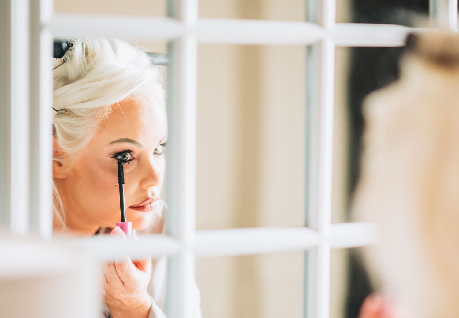 Blonde woman applying mascara while looking in a mirror during bridal makeup preparation.