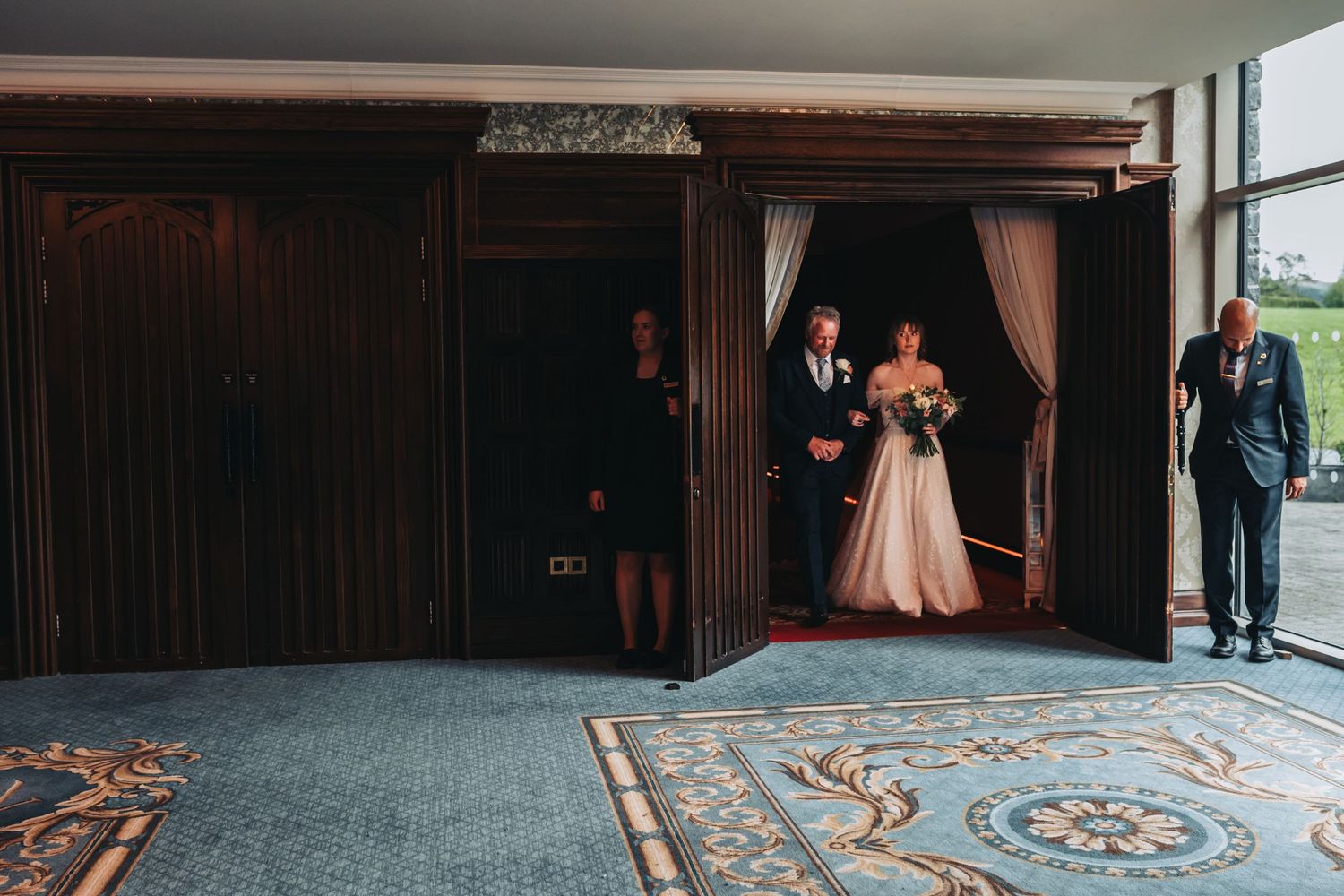 Bride and groom entering wedding venue through elegant wooden doors, with wedding party on blue carpet.