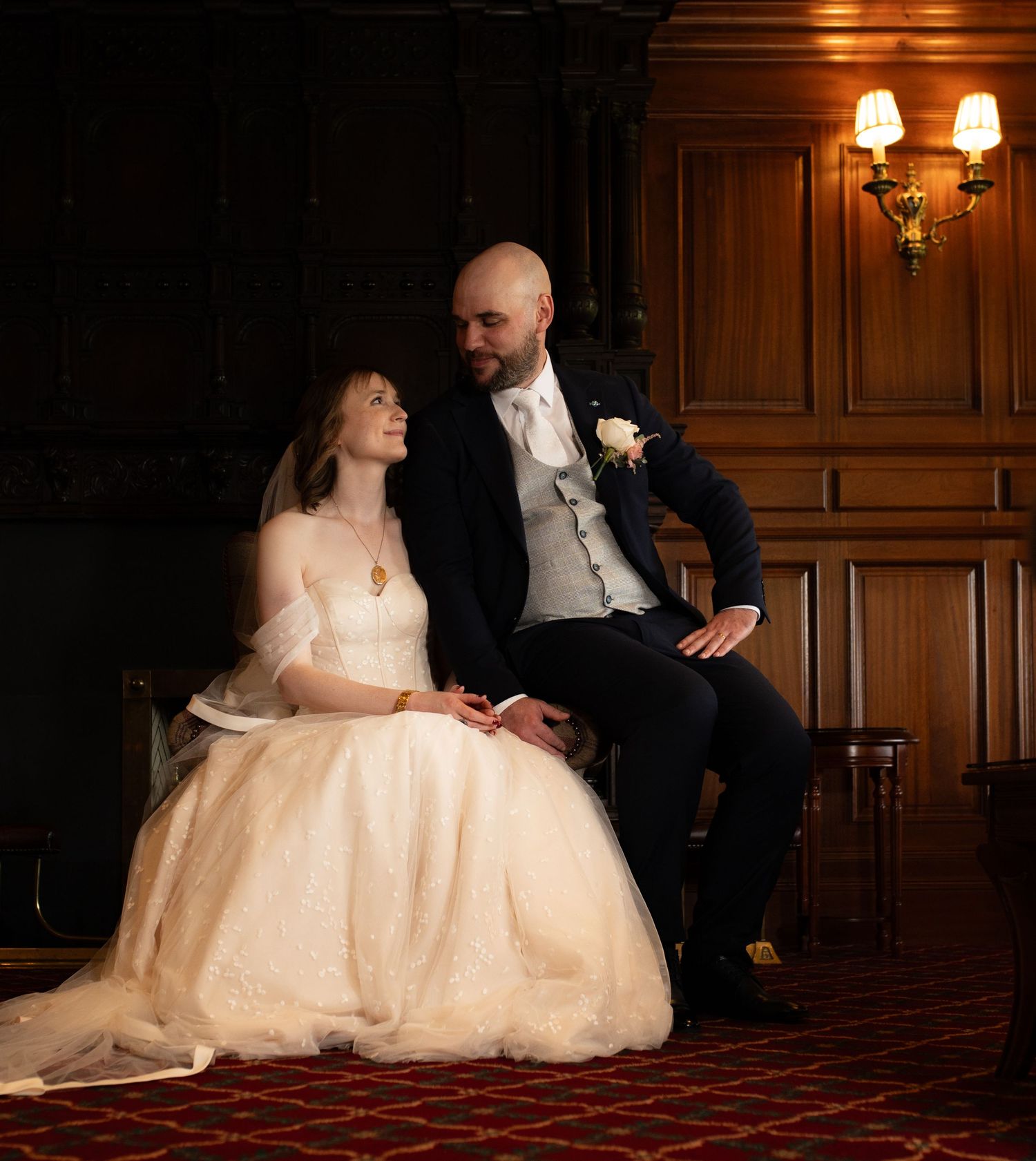 Bride in white gown and groom in navy suit share a tender moment in an elegant wood-paneled room.