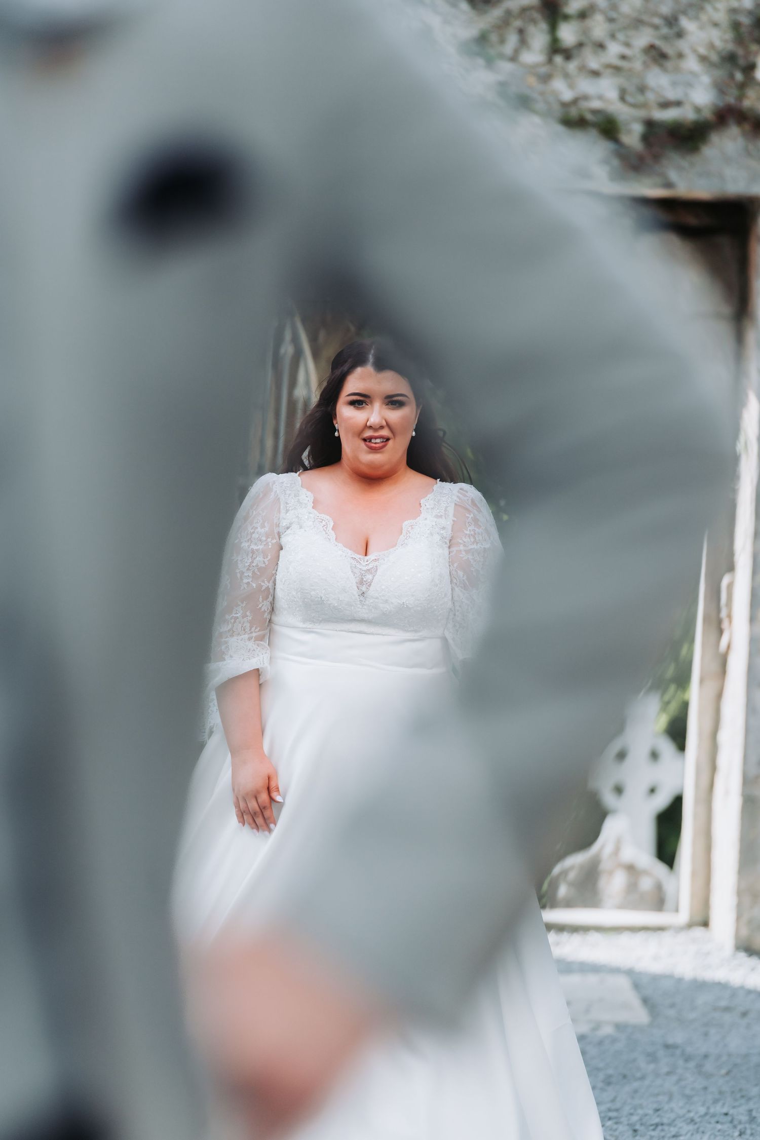 Bride in white lace wedding gown photographed through creative blurred foreground elements.