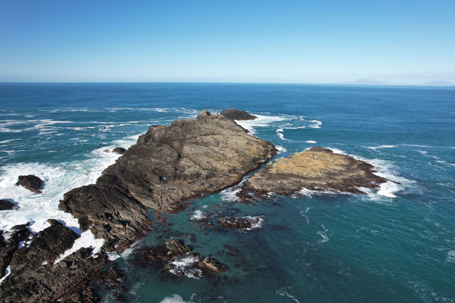 Aerial view of rocky coastal islands surrounded by turquoise ocean waves under a clear blue sky.