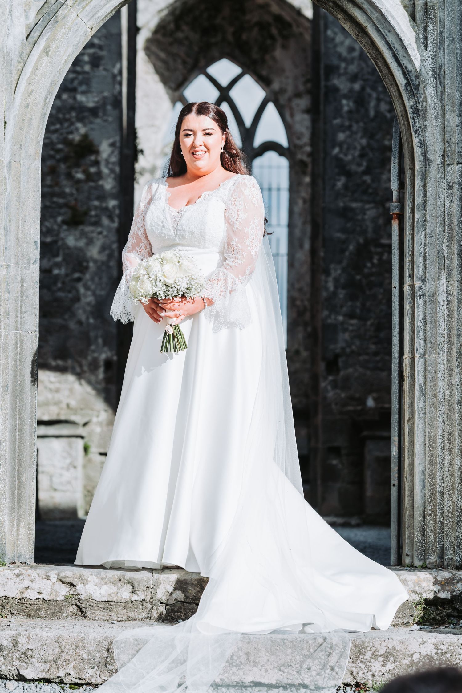Bride in white lace long-sleeve gown holding bouquet, standing in stone archway of historic building.