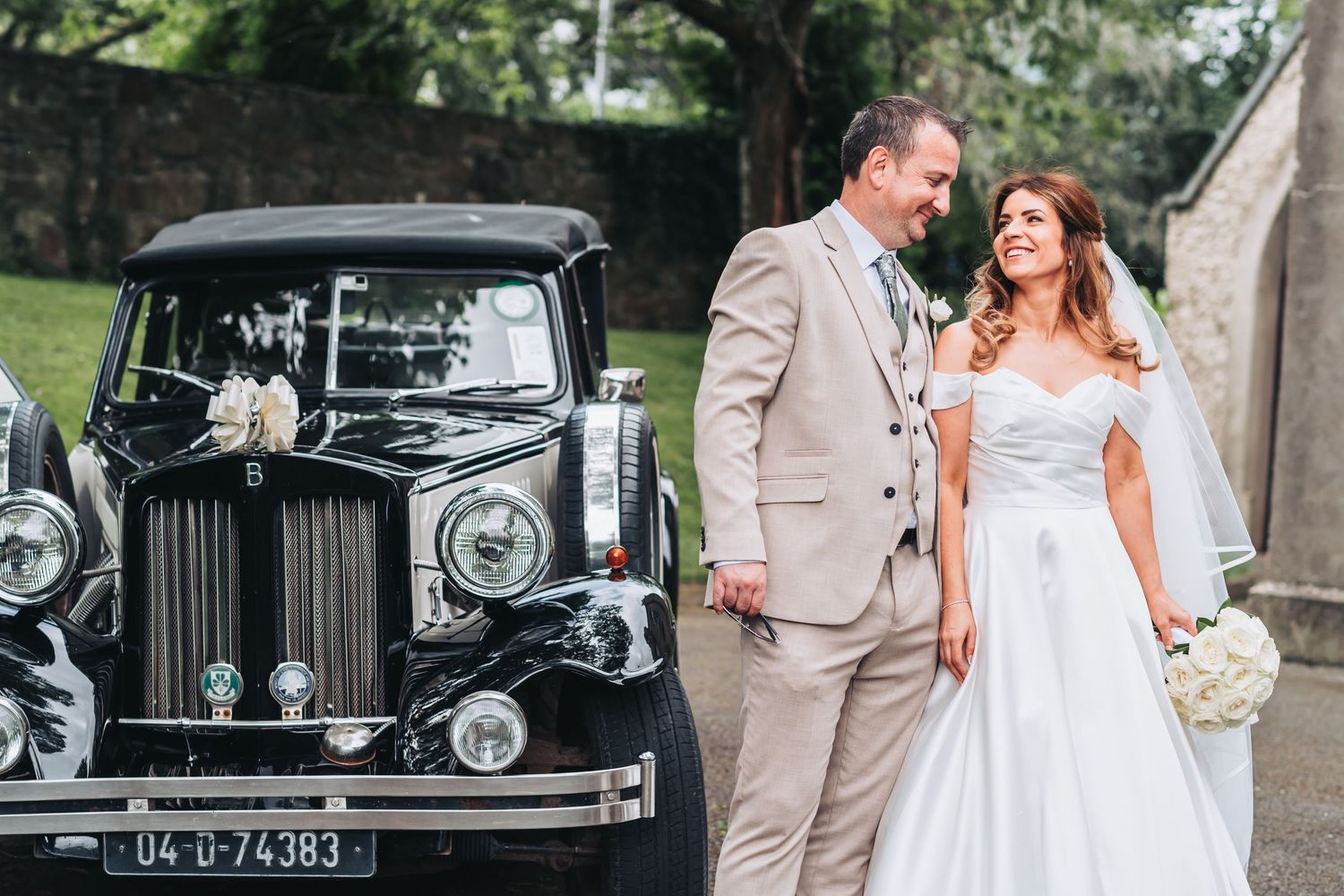 A smiling bride and groom stand beside a classic black vintage wedding car on their special day.