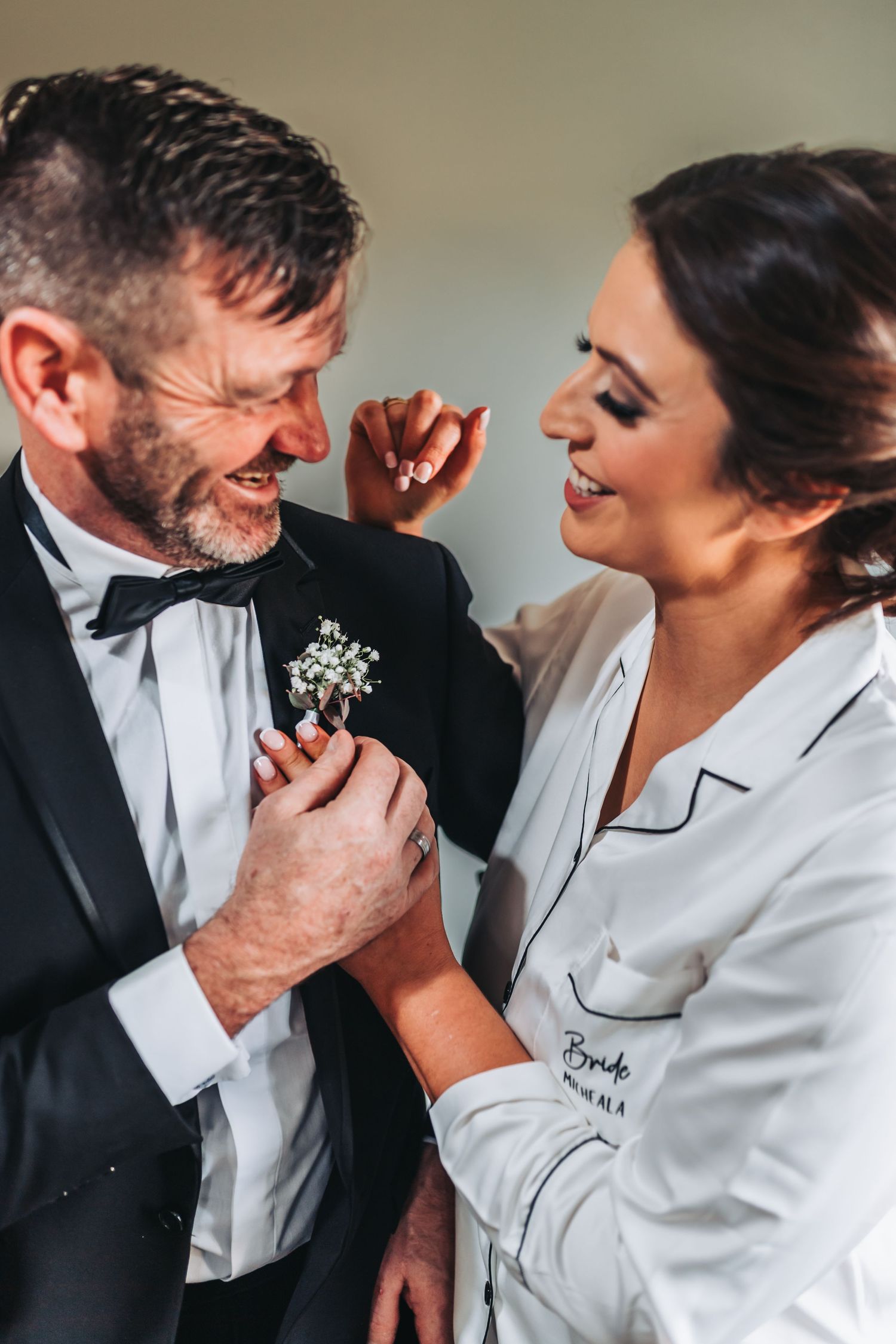 Bride in white robe pins boutonniere on groom in black tuxedo, both smiling joyfully during wedding preparations.