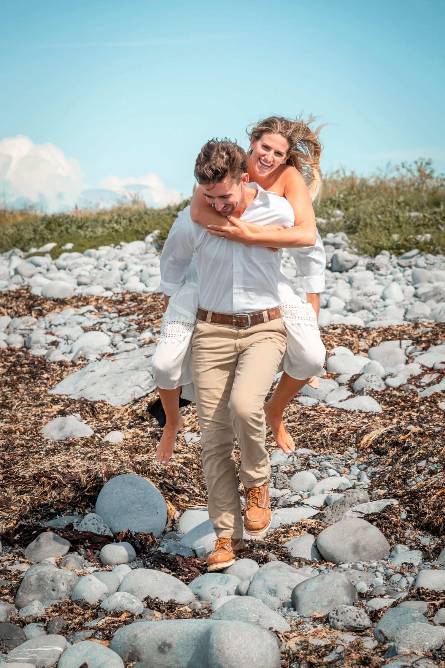 A playful couple shares a piggyback ride moment on a rocky beach shore.