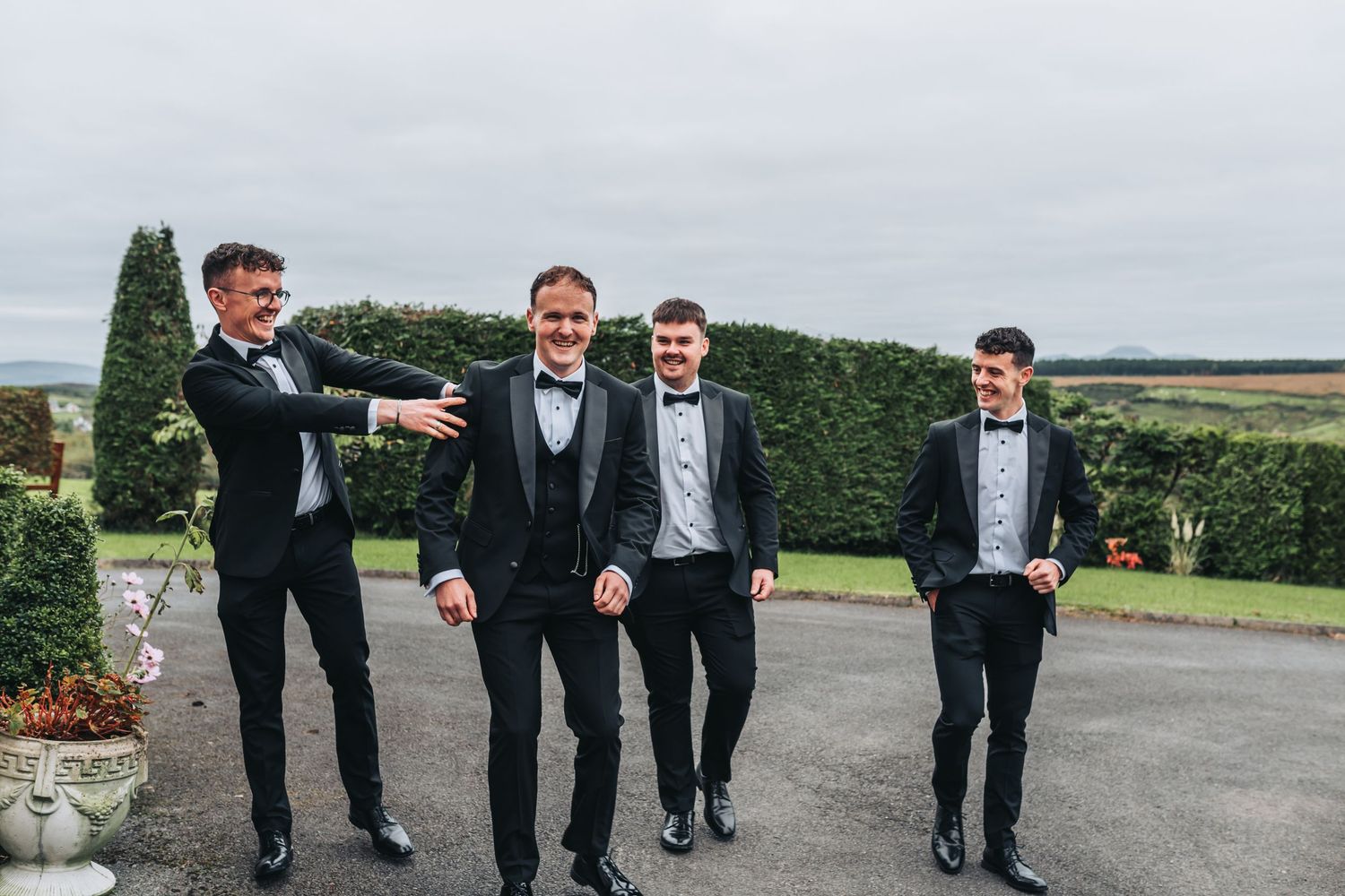 Groomsmen in black tuxedos posing casually outdoors against a hedge backdrop.