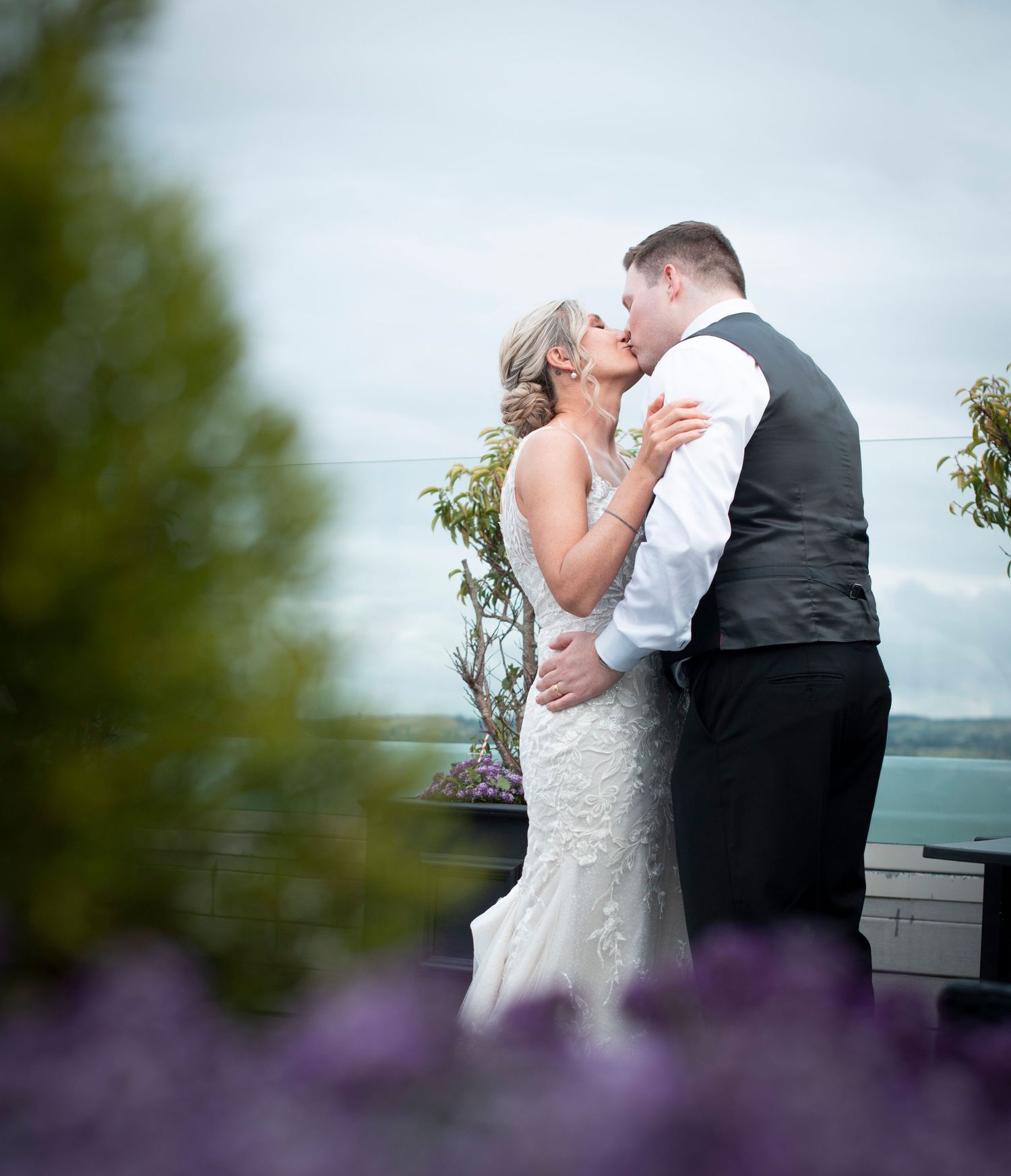 A couple embraces in formal attire with purple flowers in the foreground.
