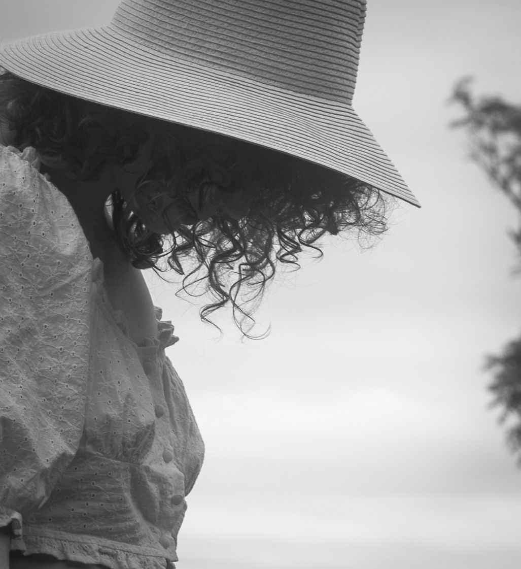Black and white photo of a woman with curly hair wearing a wide-brim striped sun hat, looking down outdoors.