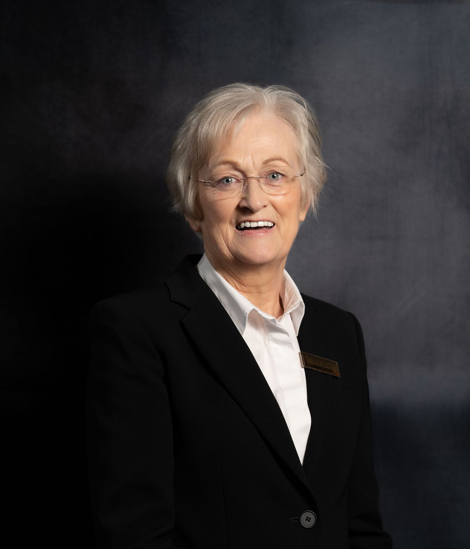 Smiling elderly woman with gray hair and glasses wearing a black blazer and white shirt against a dark background.
