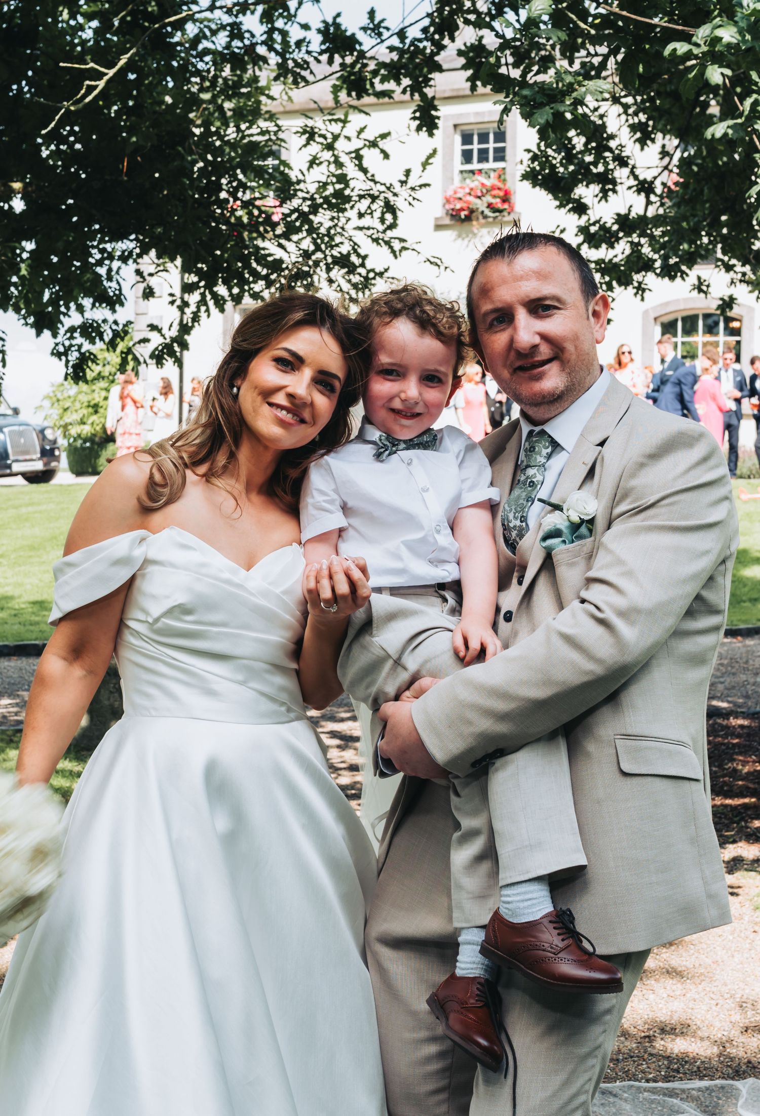 Bride and groom in beige suit hold young boy in white outfit at outdoor wedding ceremony.
