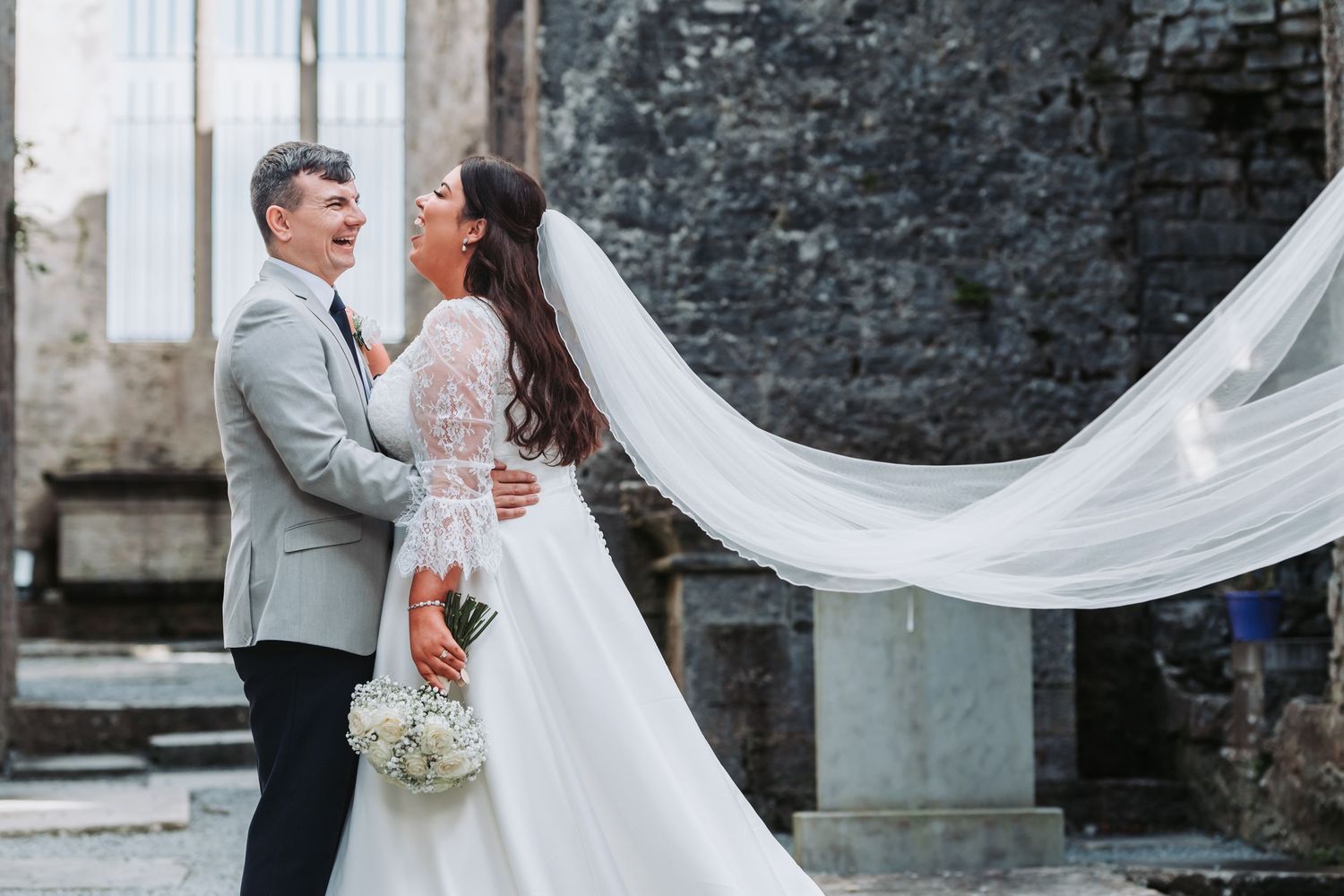 Romantic newlyweds embracing with flowing veil against rustic stone wall backdrop.