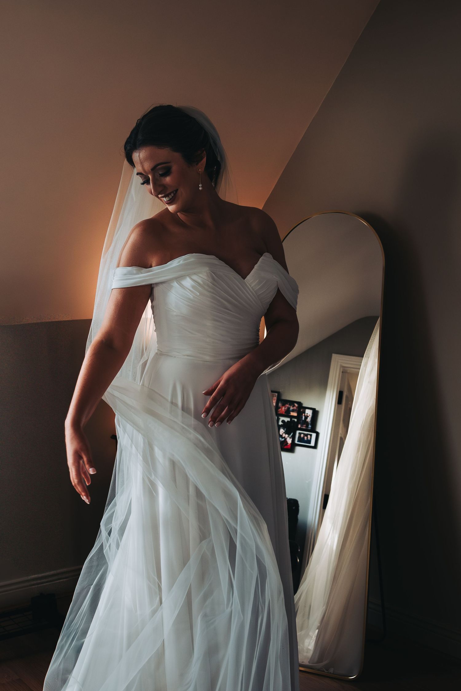 Elegant bride in flowing off-shoulder white tulle gown standing by arched mirror in soft light.