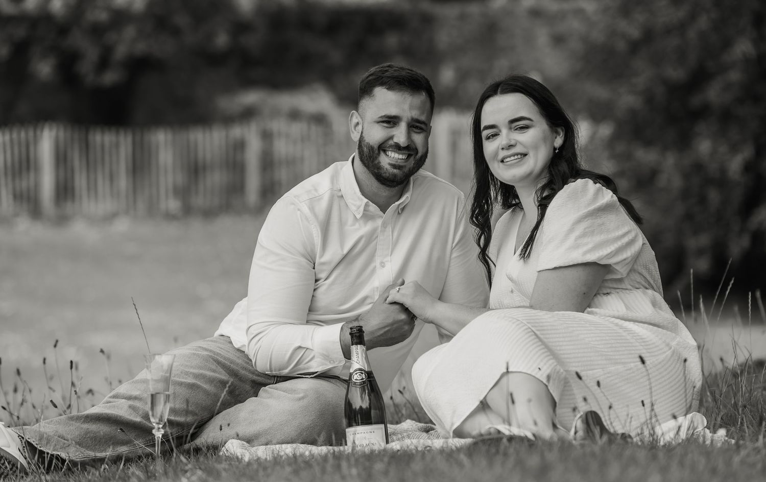 Happy couple sitting on grass enjoying a picnic with wine bottle in black and white photo.