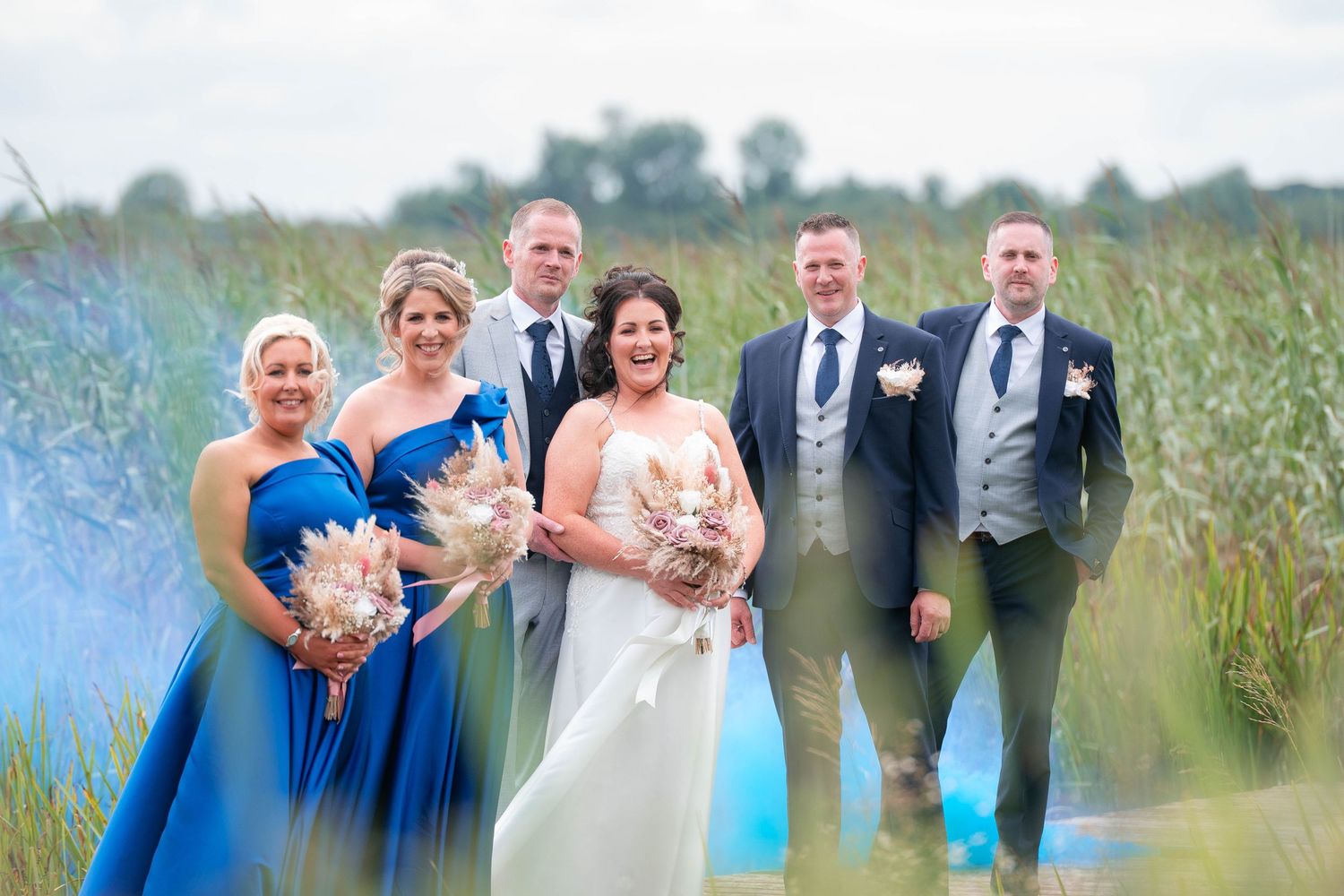 Wedding party posing outdoors near tall grass, bride in white gown with bridesmaids in blue dresses and groomsmen in suits.