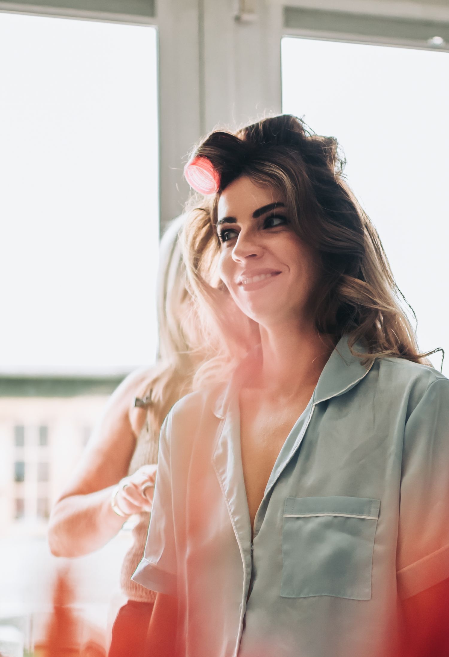 Woman with hair rollers smiling in bright room wearing light blue shirt, getting ready for an event.