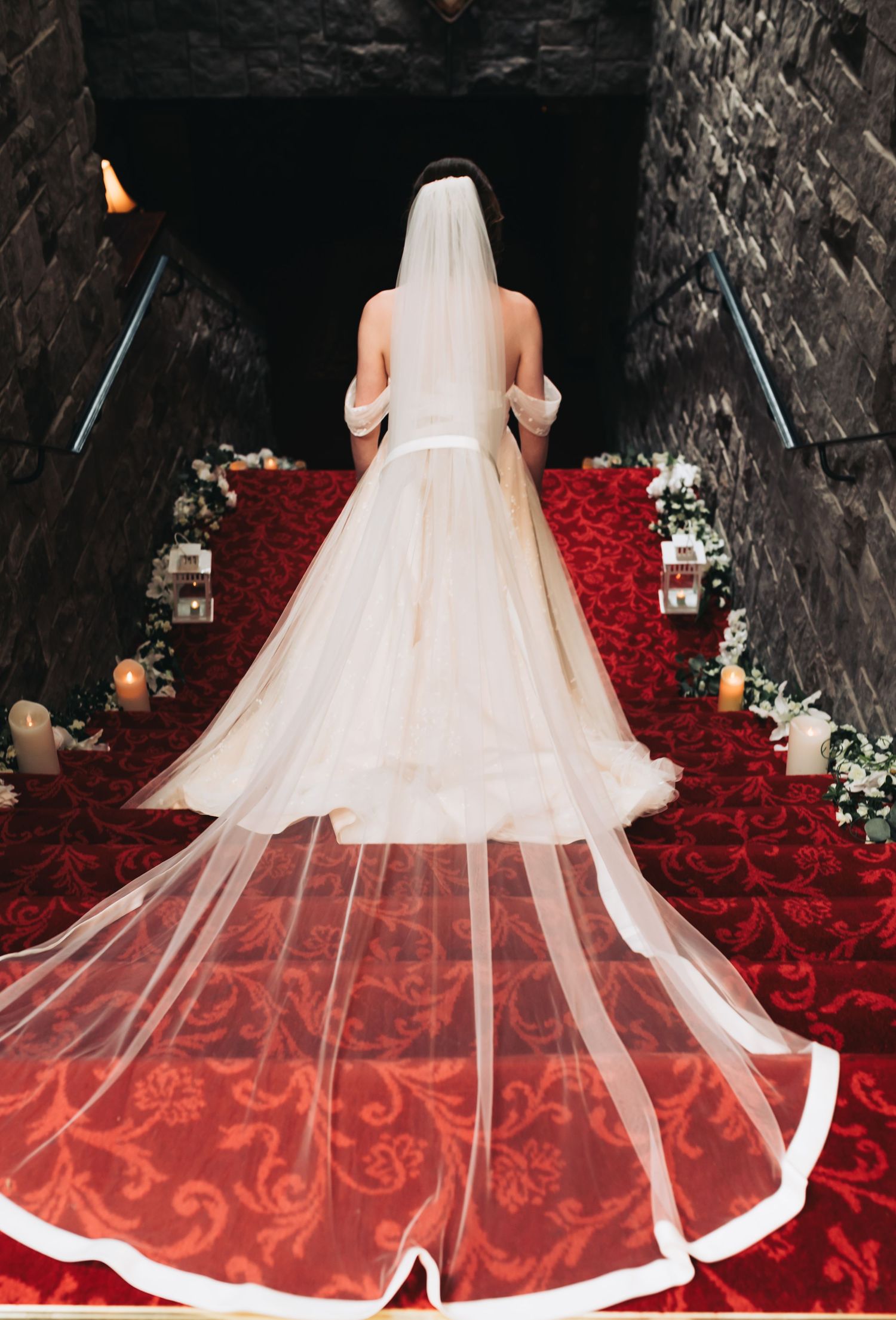Bride in off-shoulder white ball gown with cathedral veil descending red rose petal staircase.