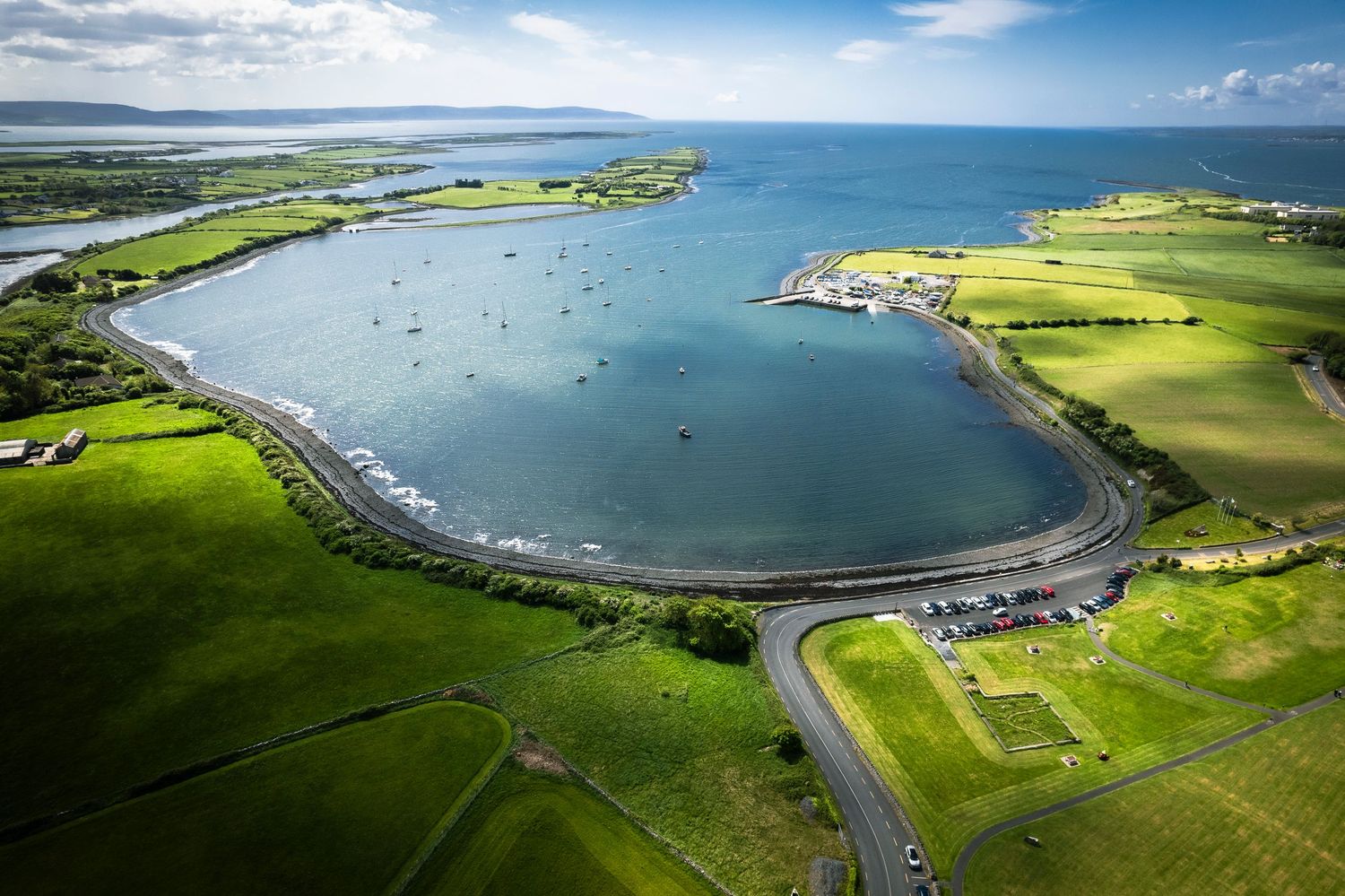 Aerial view of a calm circular harbor surrounded by green Irish farmland and blue sea.