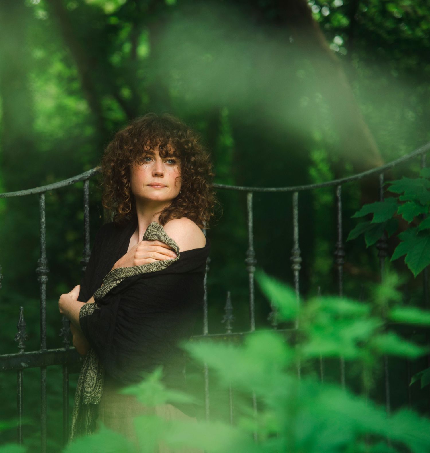Curly-haired woman in black wrap leaning on iron gate surrounded by green foliage.