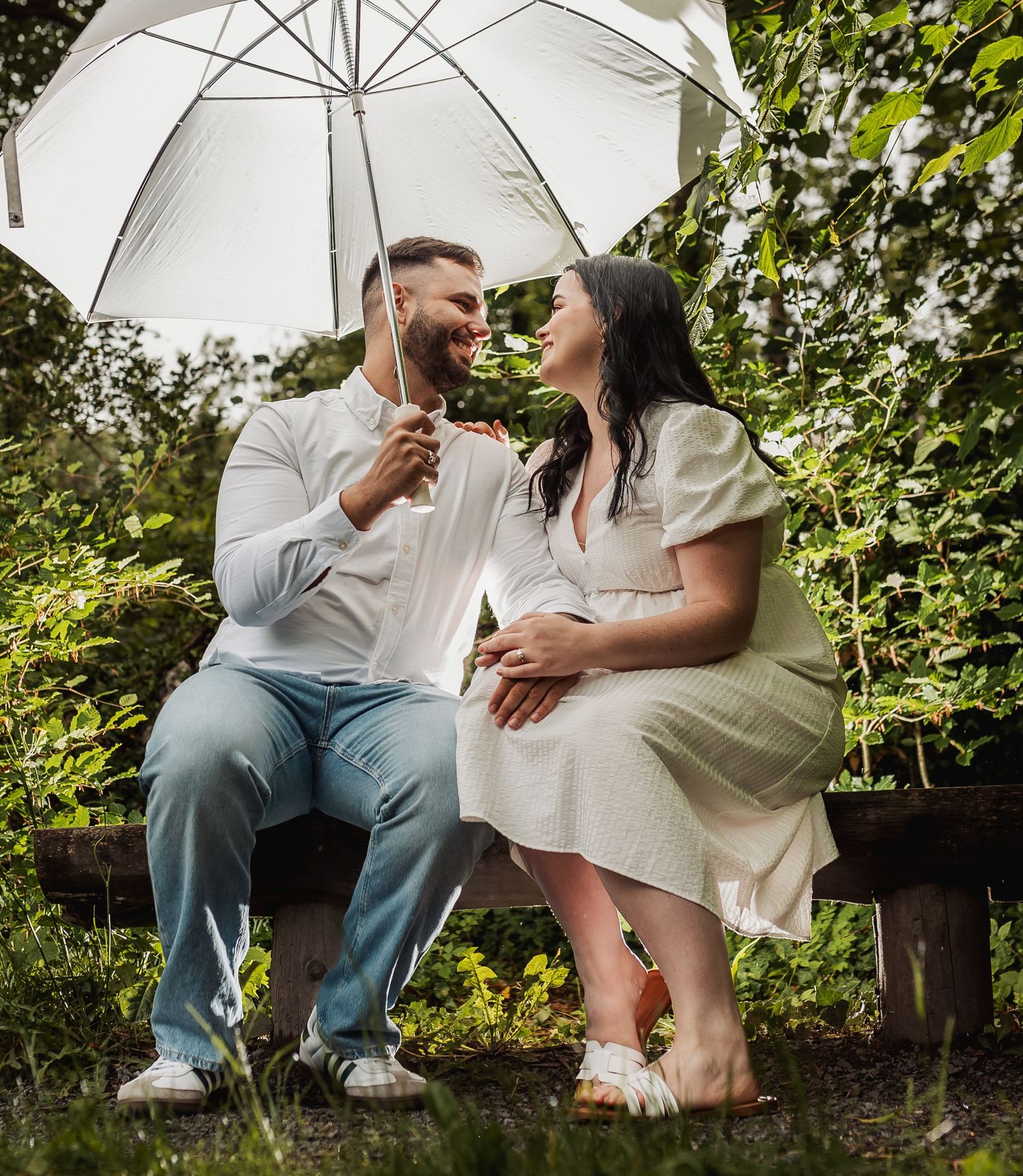 Couple sitting on a bench under a white umbrella, smiling at each other, surrounded by lush green foliage.