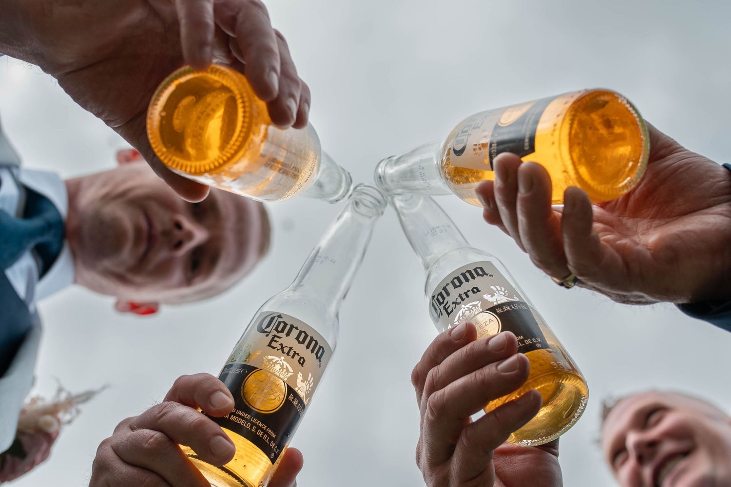 Four friends toasting with Corona Extra beer bottles outdoors, viewed from below against a cloudy sky.