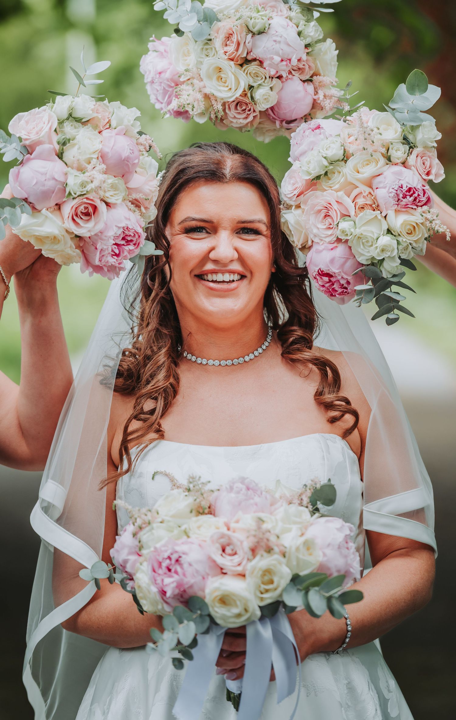 Smiling bride in white gown and veil holds pink and white peony bouquets, surrounded by floral arrangements outdoors.
