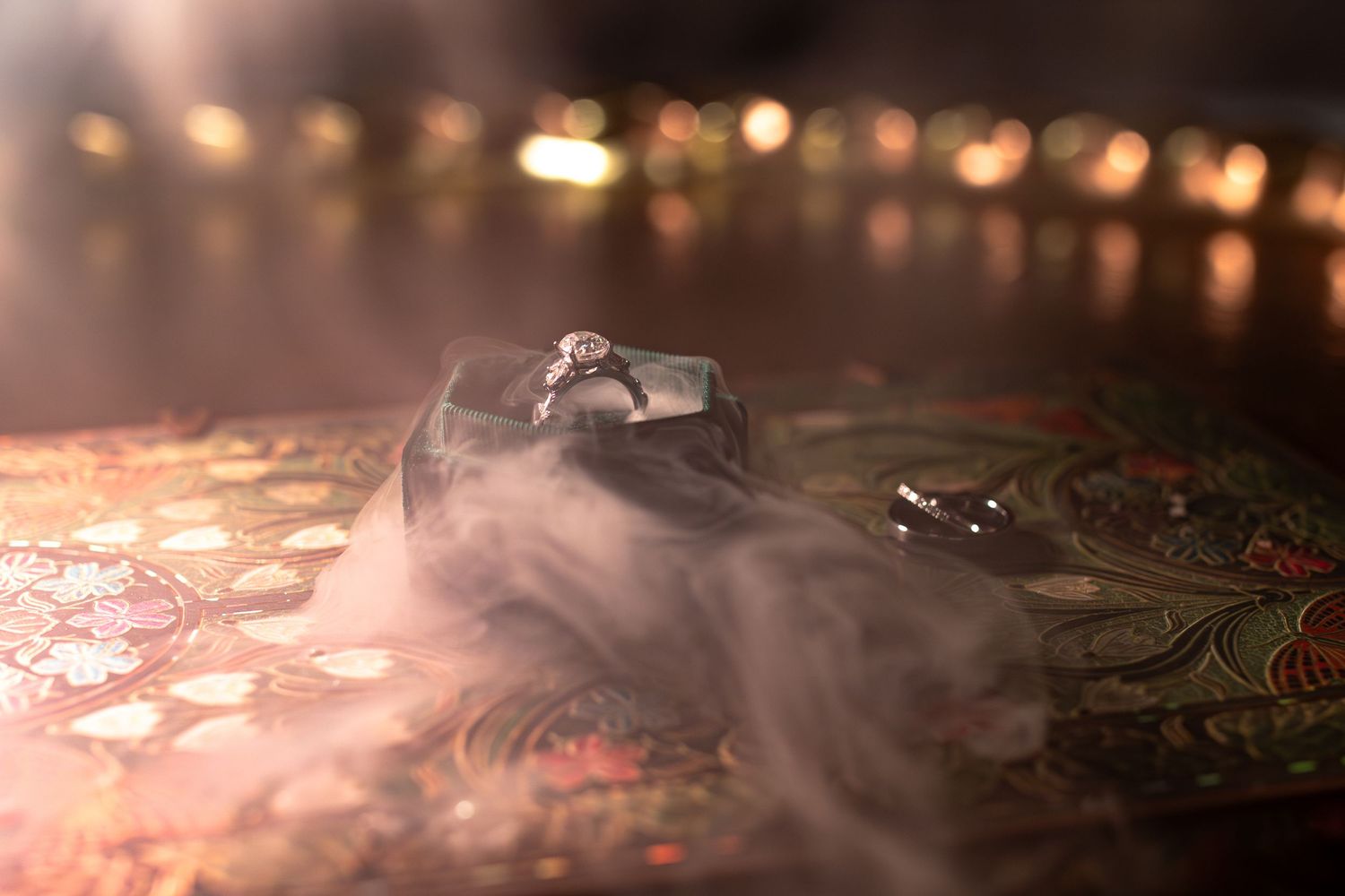 Diamond engagement ring in a black box surrounded by dramatic smoke on an ornate floral surface with bokeh lights.