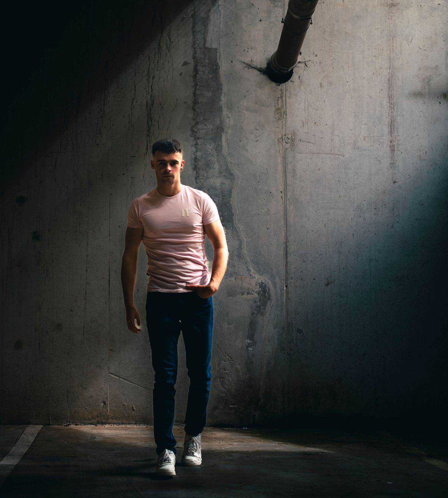 A muscular man in a pink t-shirt and blue jeans stands confidently in a dramatic concrete space with moody lighting.