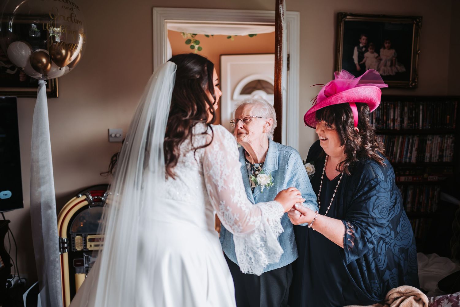 Bride in white gown sharing a tender moment with elderly grandmother and female guest in pink hat.