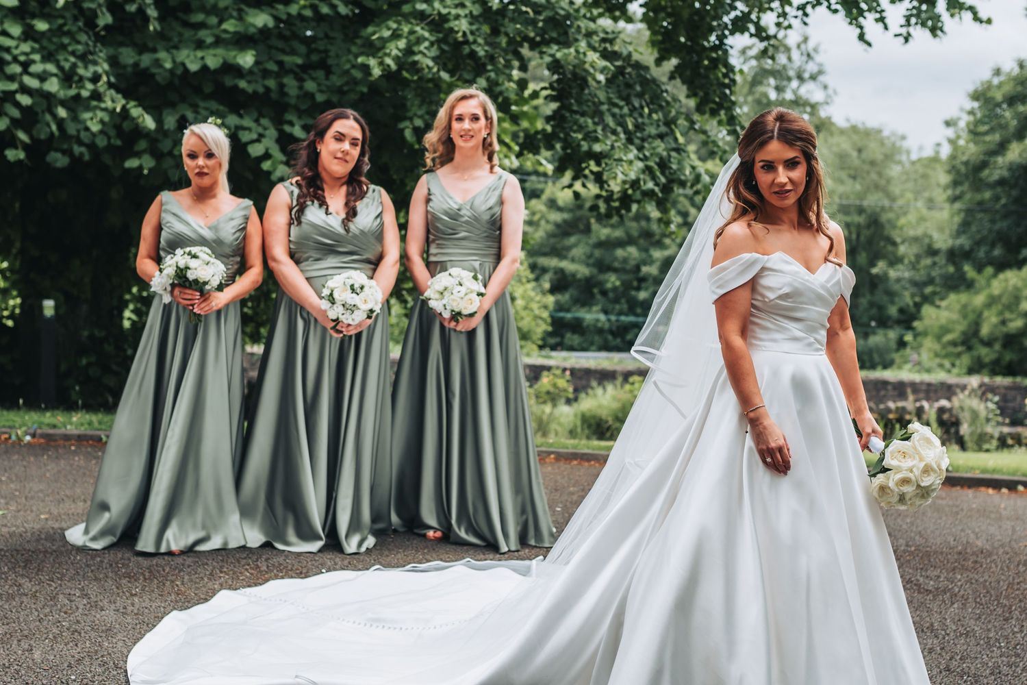 Bride in white ball gown with three bridesmaids in sage green dresses outdoors.