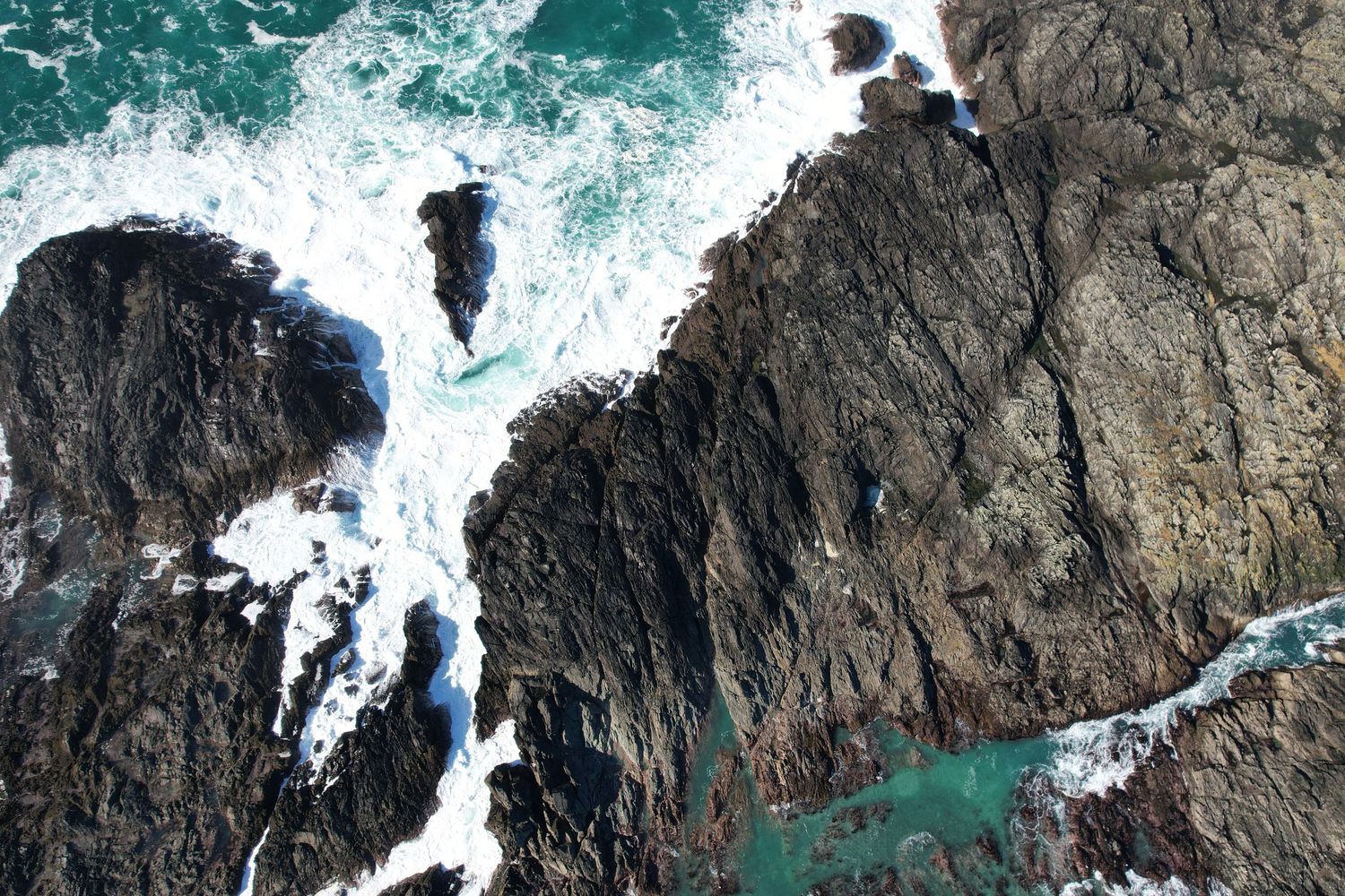 Aerial view of rugged rocky coastline with turquoise ocean waves crashing against dark jagged rocks.