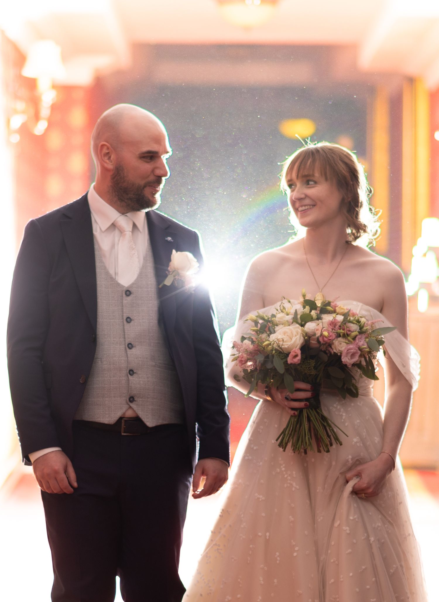 Bald groom in a navy suit and bride in a white gown holding a pink bouquet walk together at their wedding.