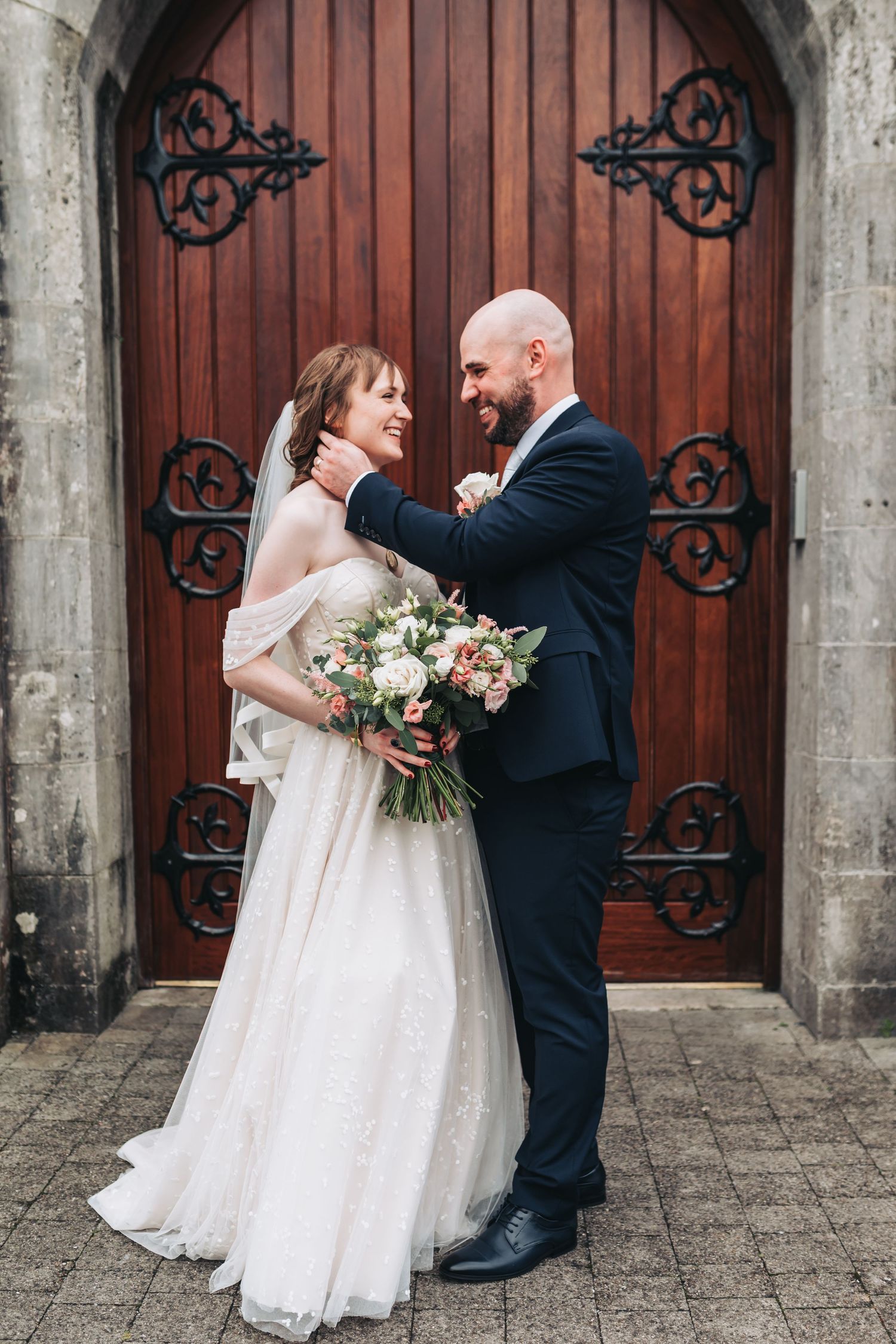 Bride and groom embrace before ornate wooden church doors, holding a lush floral bouquet.