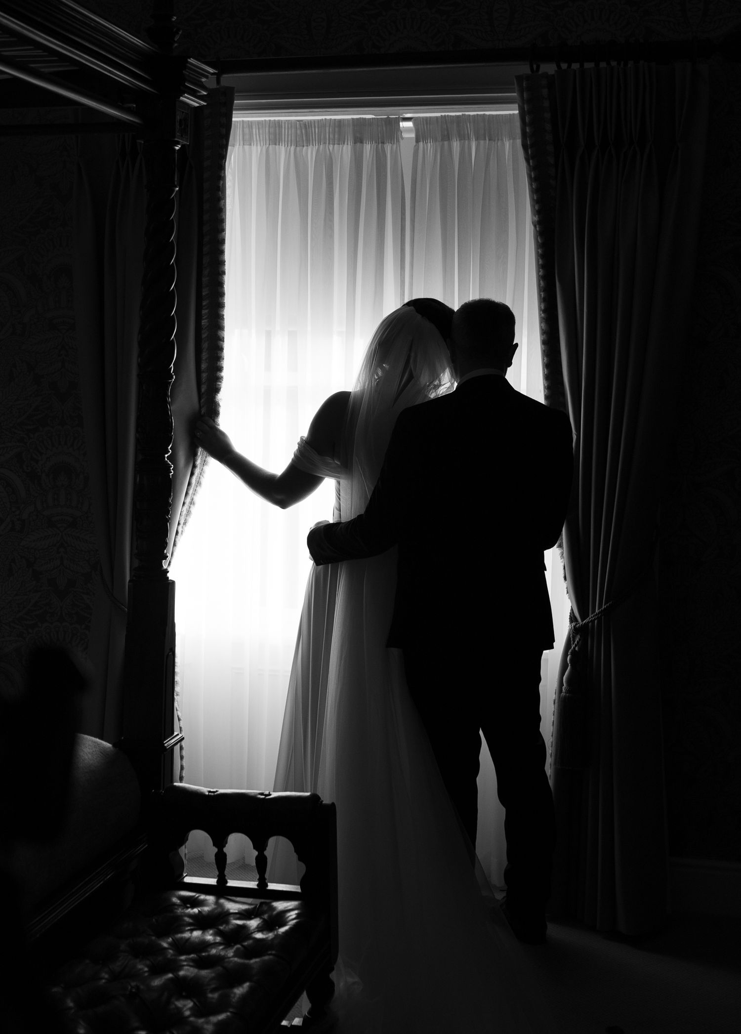 Silhouette of bride and groom embracing by a bright window in a dark elegant room on their wedding day.