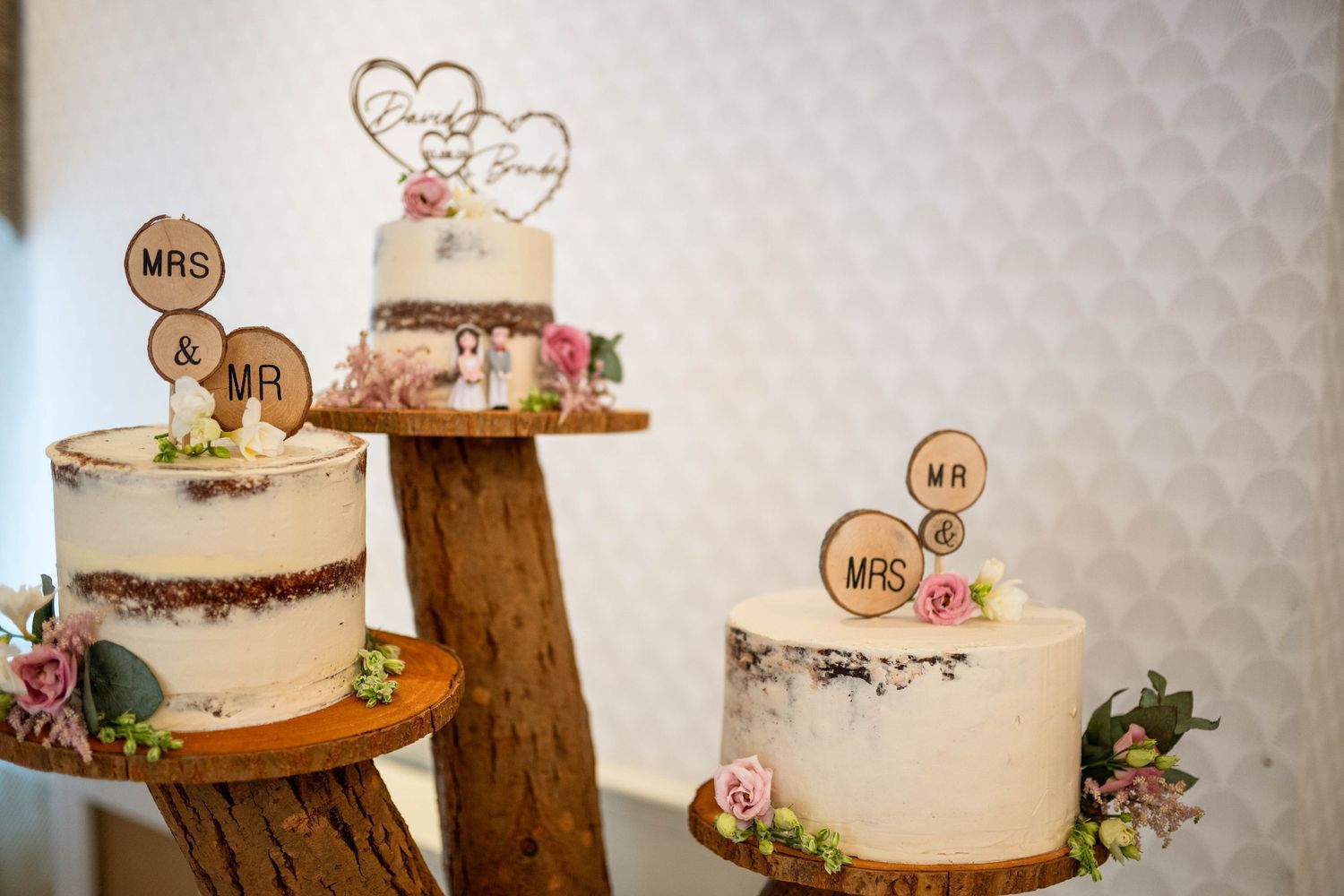 Three rustic naked wedding cakes on wooden stands decorated with Mr & Mrs toppers and pink flowers.