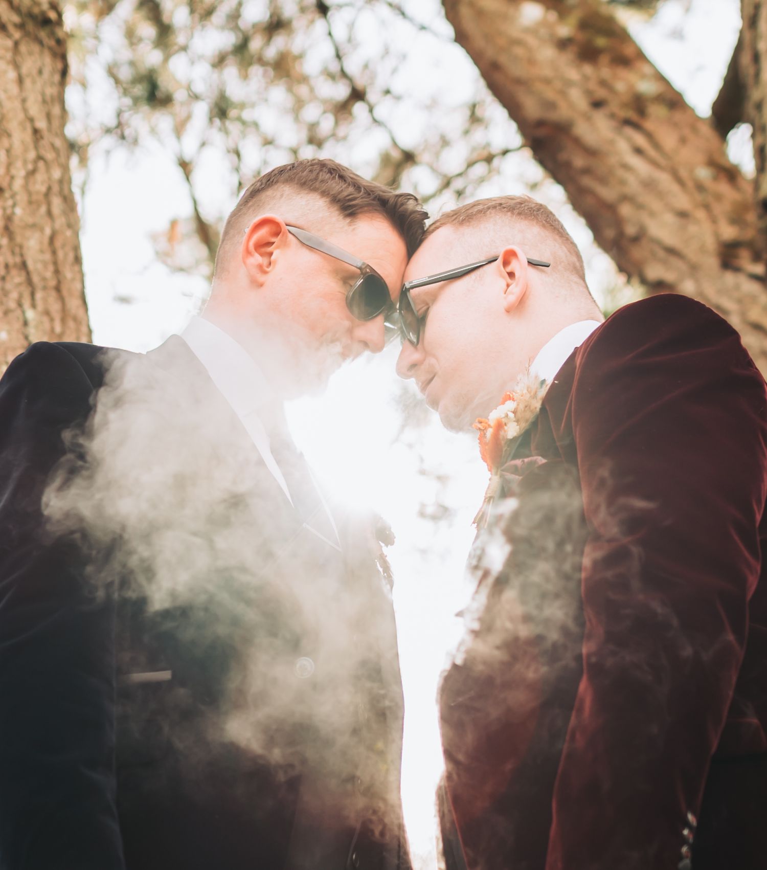 Two grooms in velvet suits touching foreheads surrounded by smoke under a tree on their wedding day.