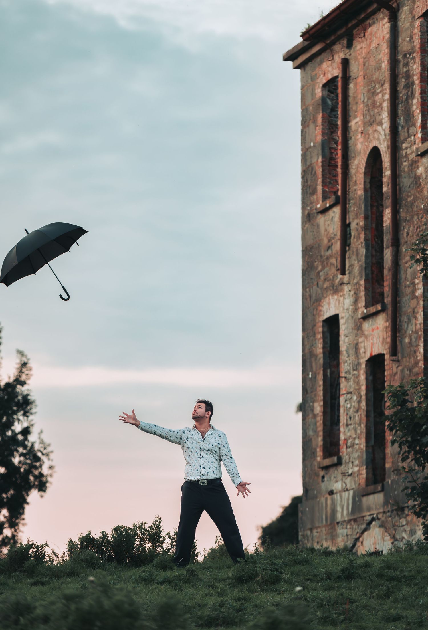 Man tossing a black umbrella into the air near a rustic brick building under a cloudy sky.