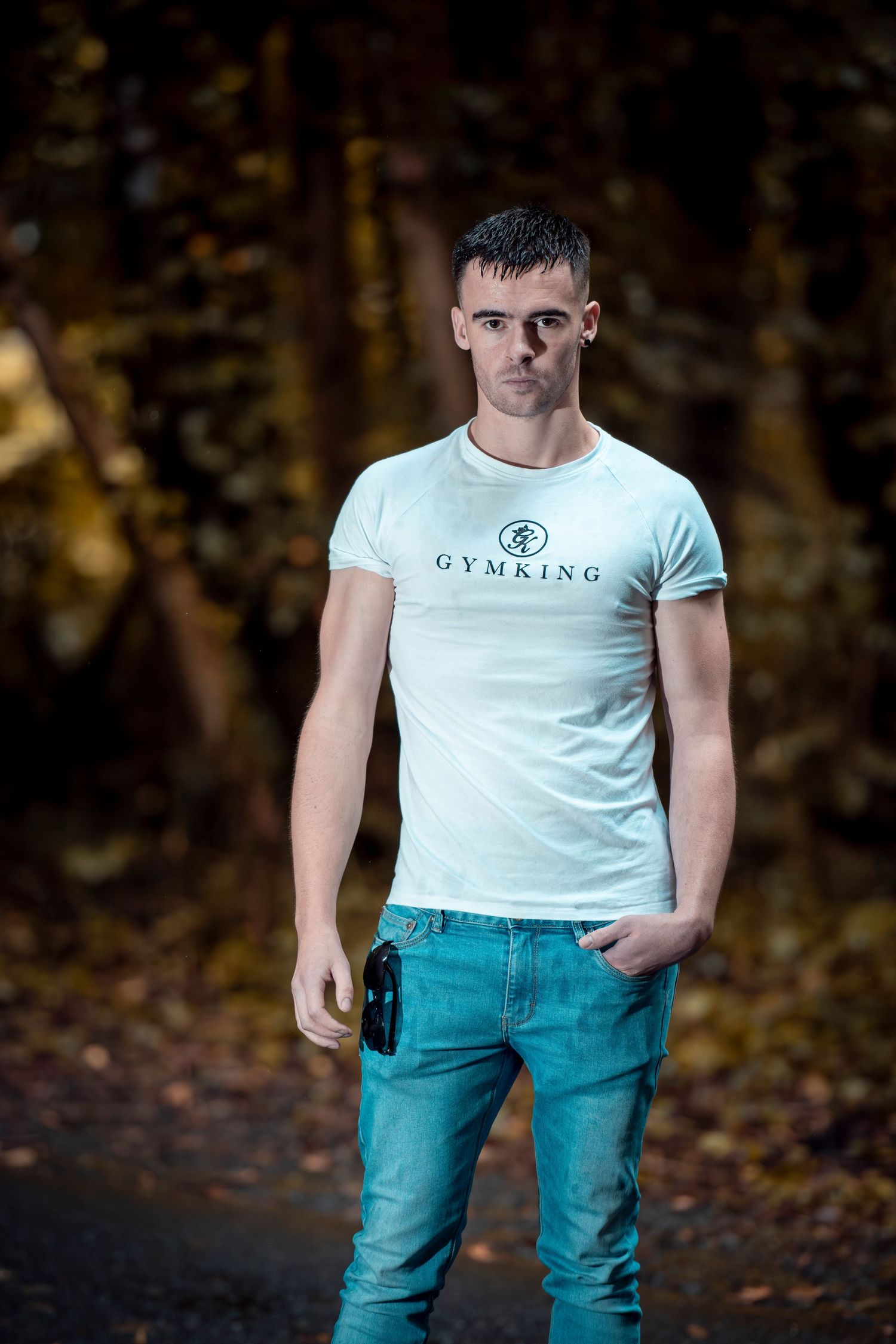 Young muscular man in white Gymking t-shirt and blue jeans standing on forest path.